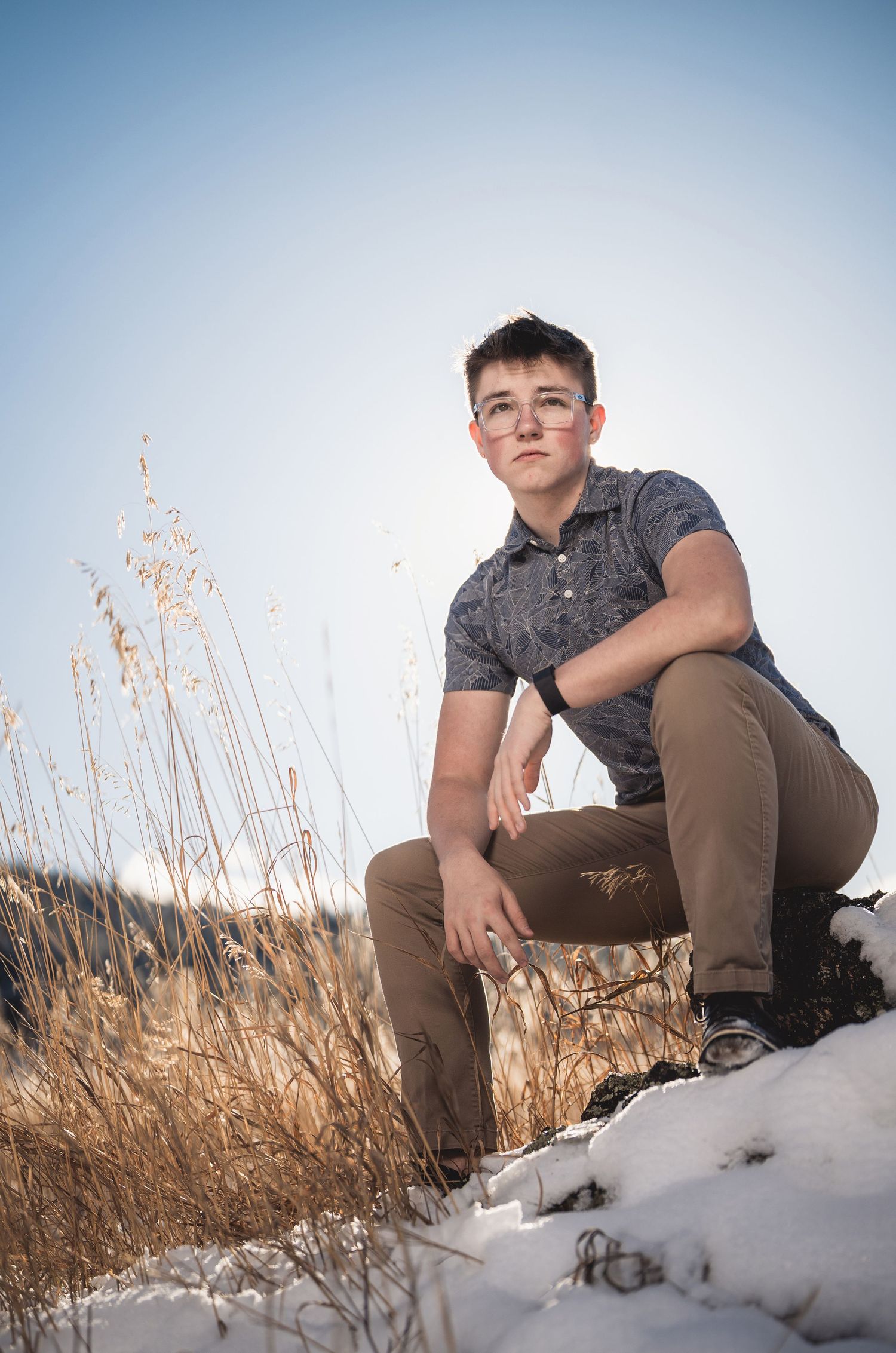 Teen boy crouching in a snowy field with dry golden grasses, wearing a gray polo and khaki pants.