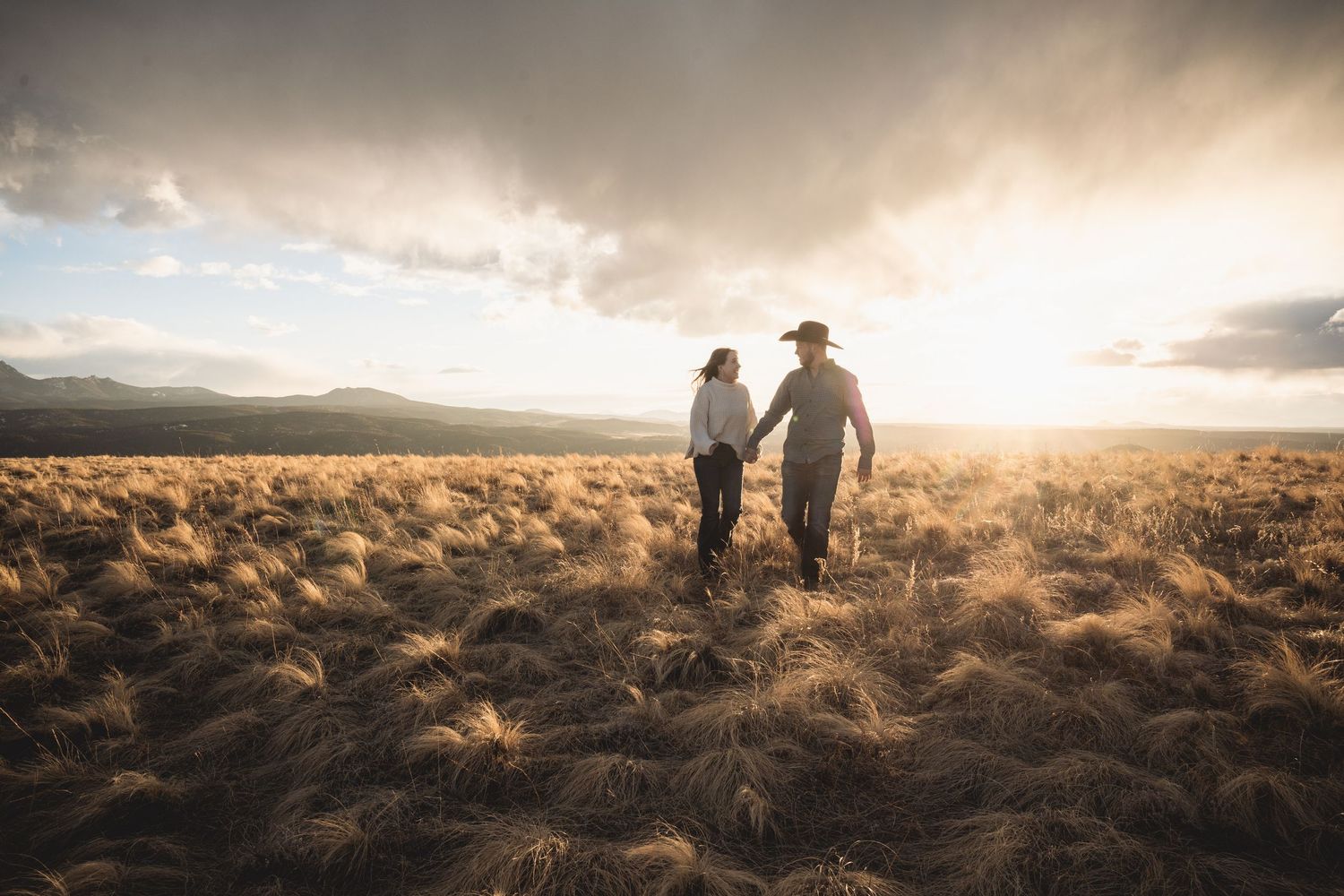 Two figures walk through golden prairie grass at sunset with mountains on horizon.