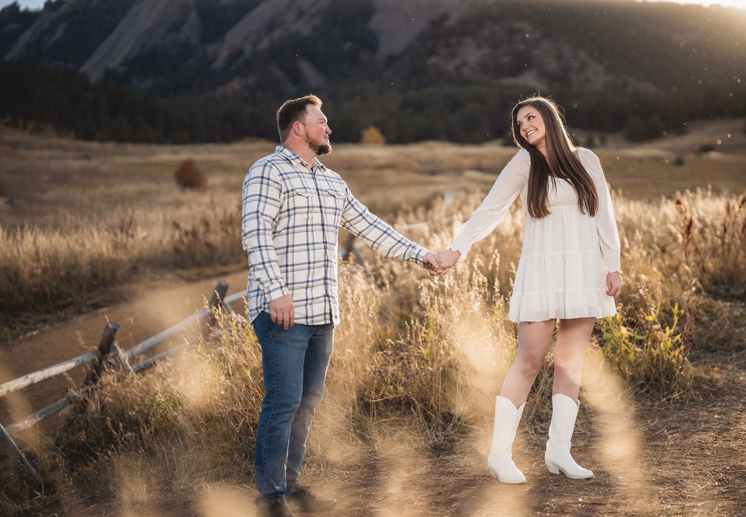Couple poses in golden field during sunset with mountain backdrop creating romantic autumn engagement photos.