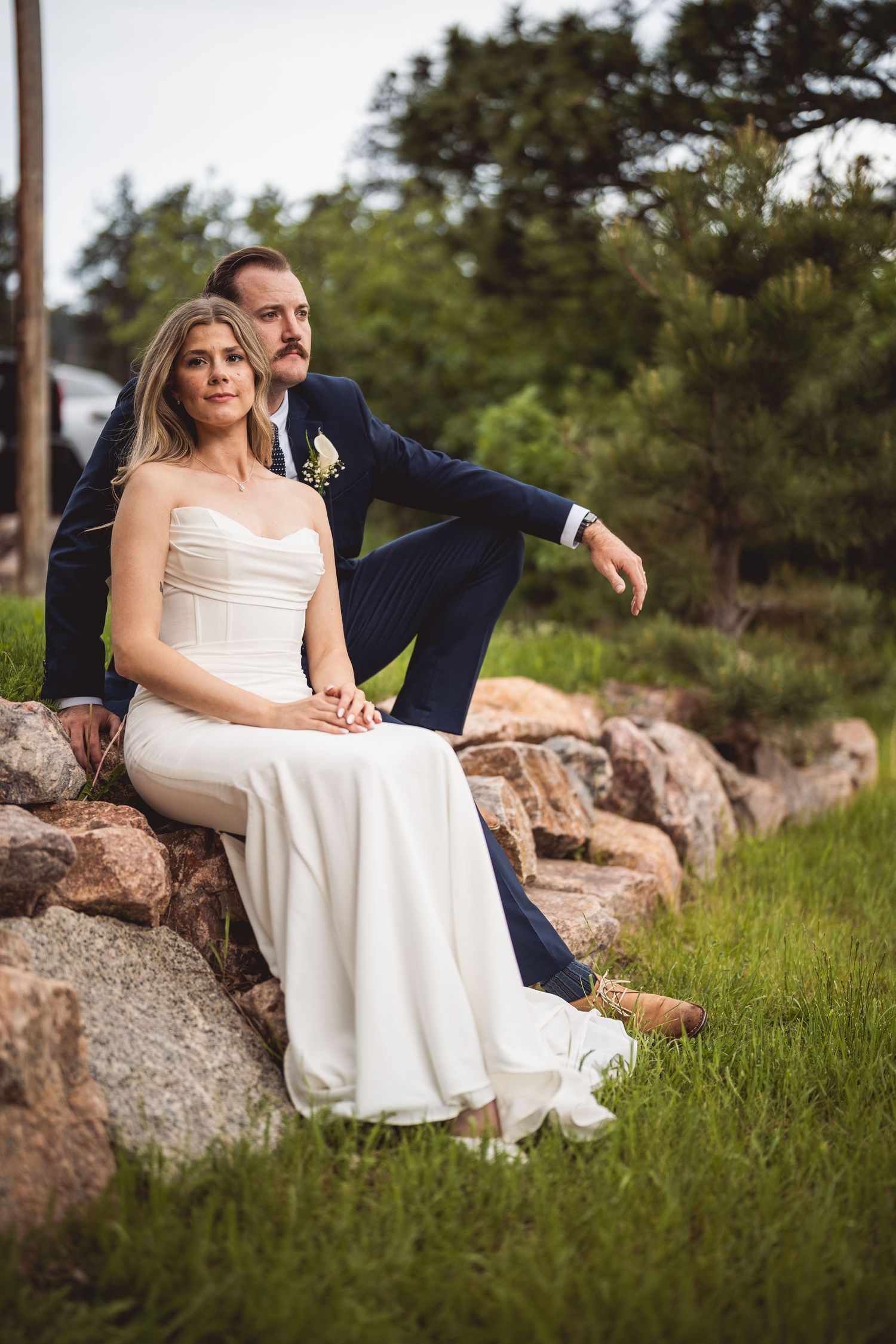 A newlywed couple poses on rocky terrain with pine trees and mountains in the background.
