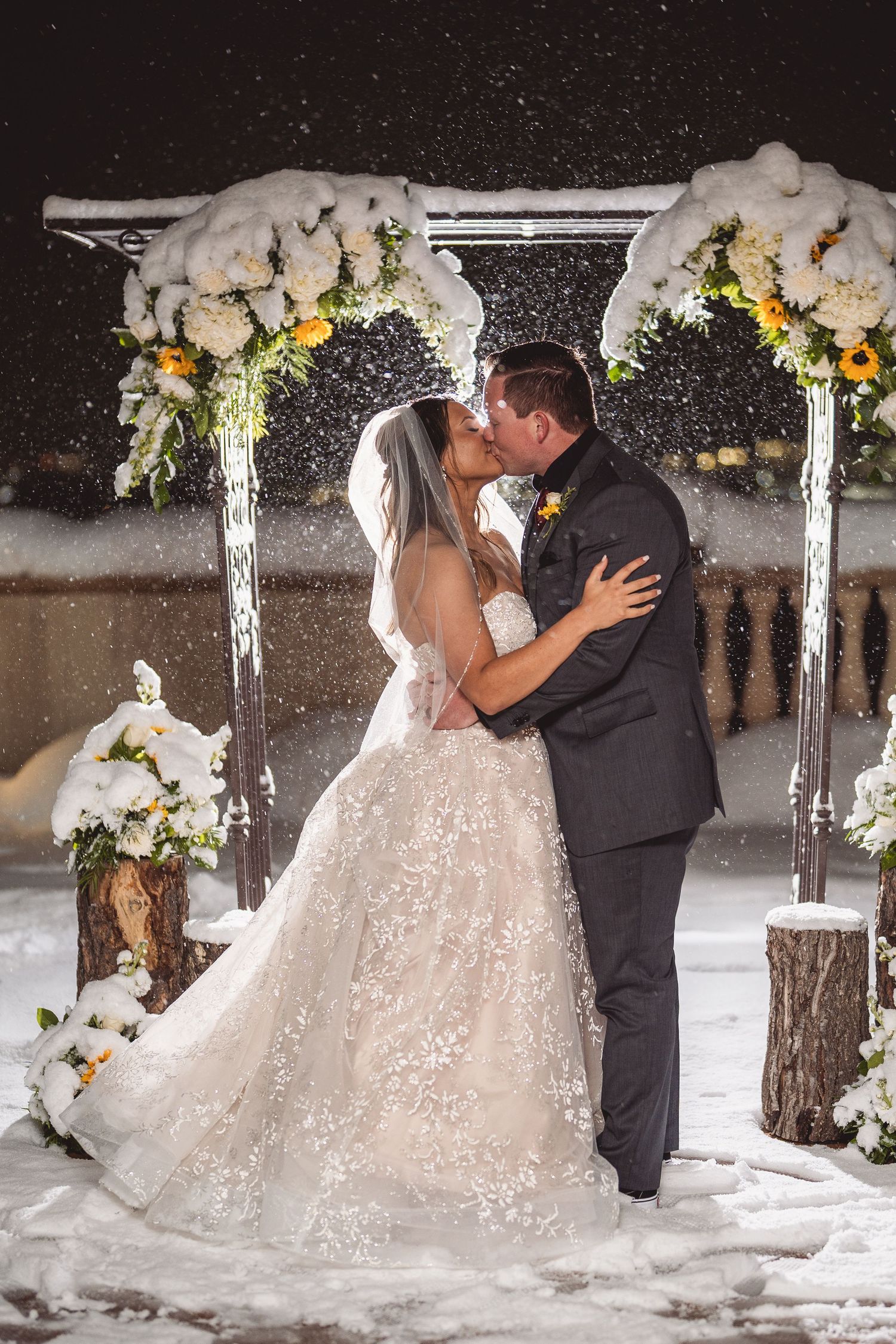 A romantic winter wedding moment with the couple sharing a kiss under a decorated arbor in falling snow.
