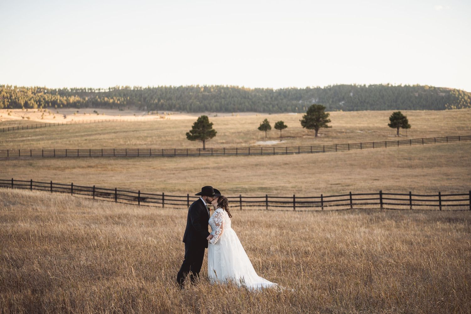 A tender moment captured in a vast countryside field with wooden fencing and scattered trees.