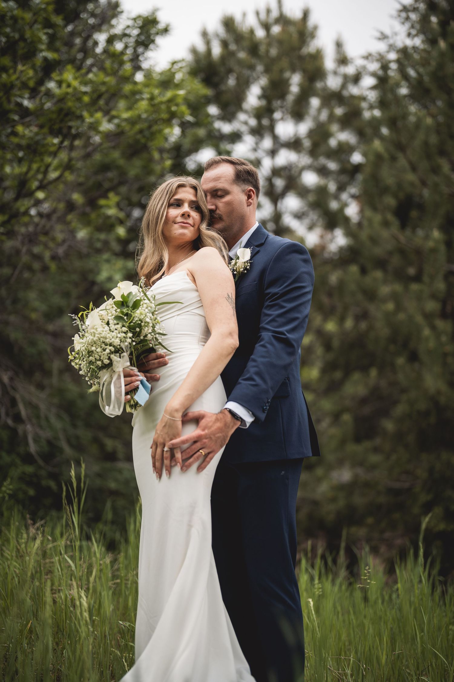 Bride in white gown holding bouquet embraced by groom in navy suit outdoors with lush green trees.