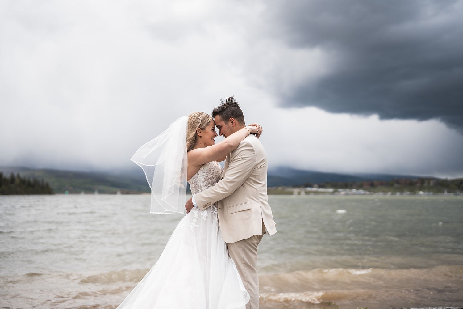 Bride and groom embrace on a lakeside beach with dramatic stormy skies, wedding veil blowing in the wind.