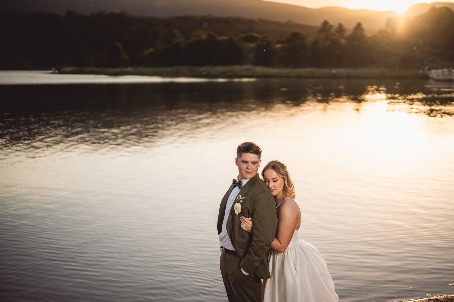 Bride and groom embrace by a serene lake at golden sunset during their romantic wedding photo session.