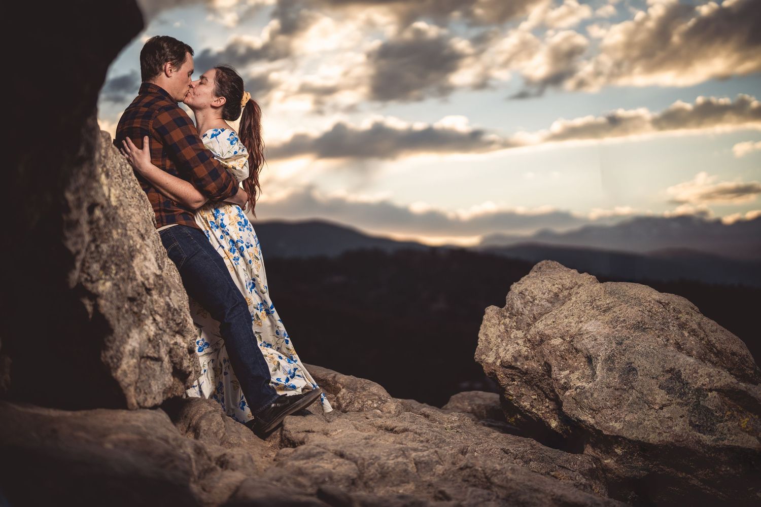 Couple kissing romantically among rocky mountain boulders at sunset with dramatic cloudy sky backdrop.
