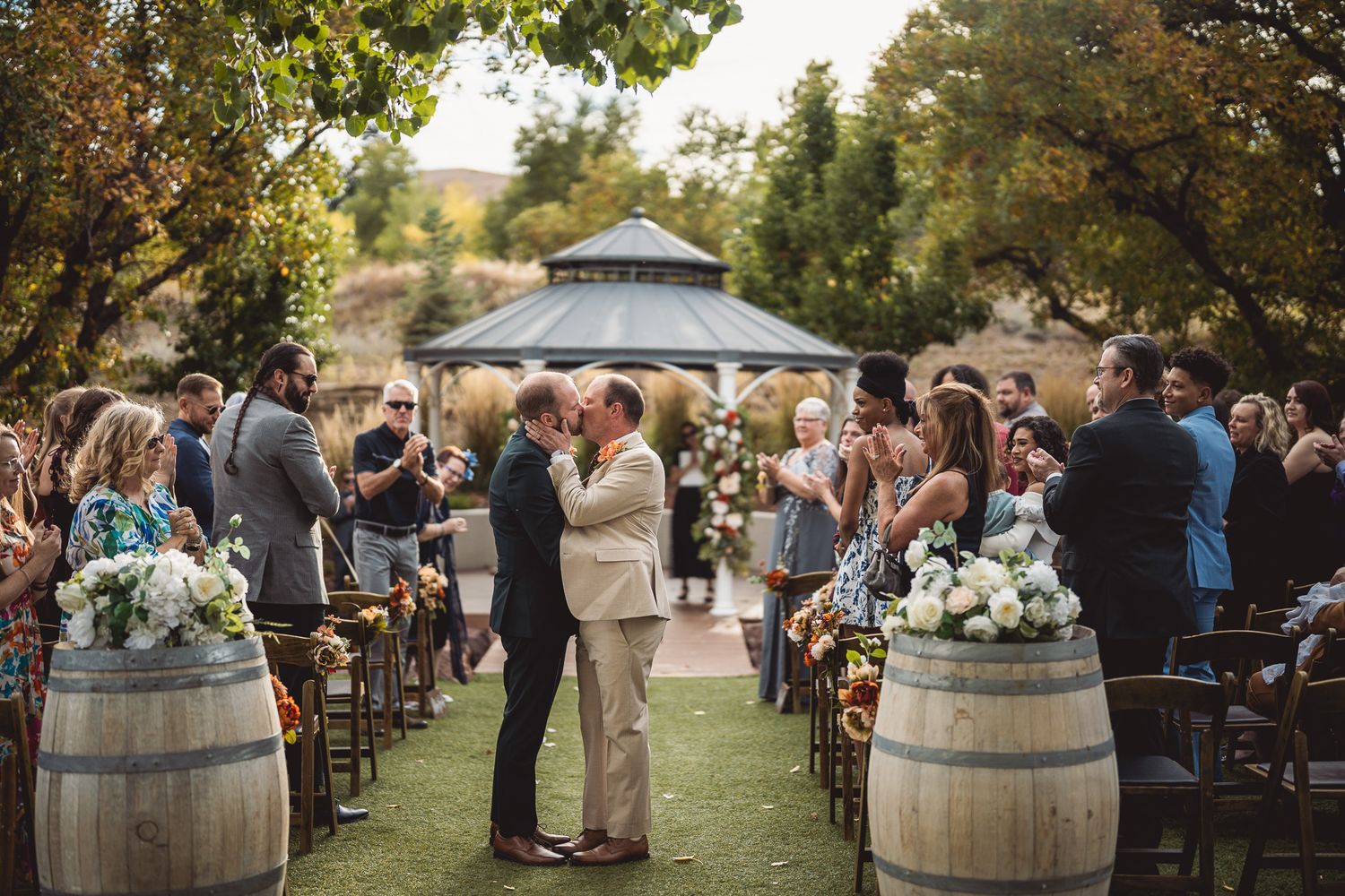 Two grooms share first kiss at outdoor winery wedding ceremony with barrel aisle decor and gazebo backdrop.