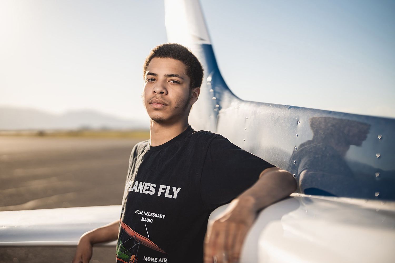 Young man in black t-shirt leaning on small aircraft wing at airfield during golden hour sunset.