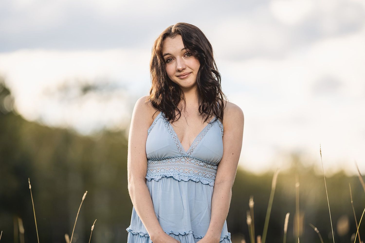 Young woman in a light blue sundress standing in a sunlit field with tall grass and trees in the background.