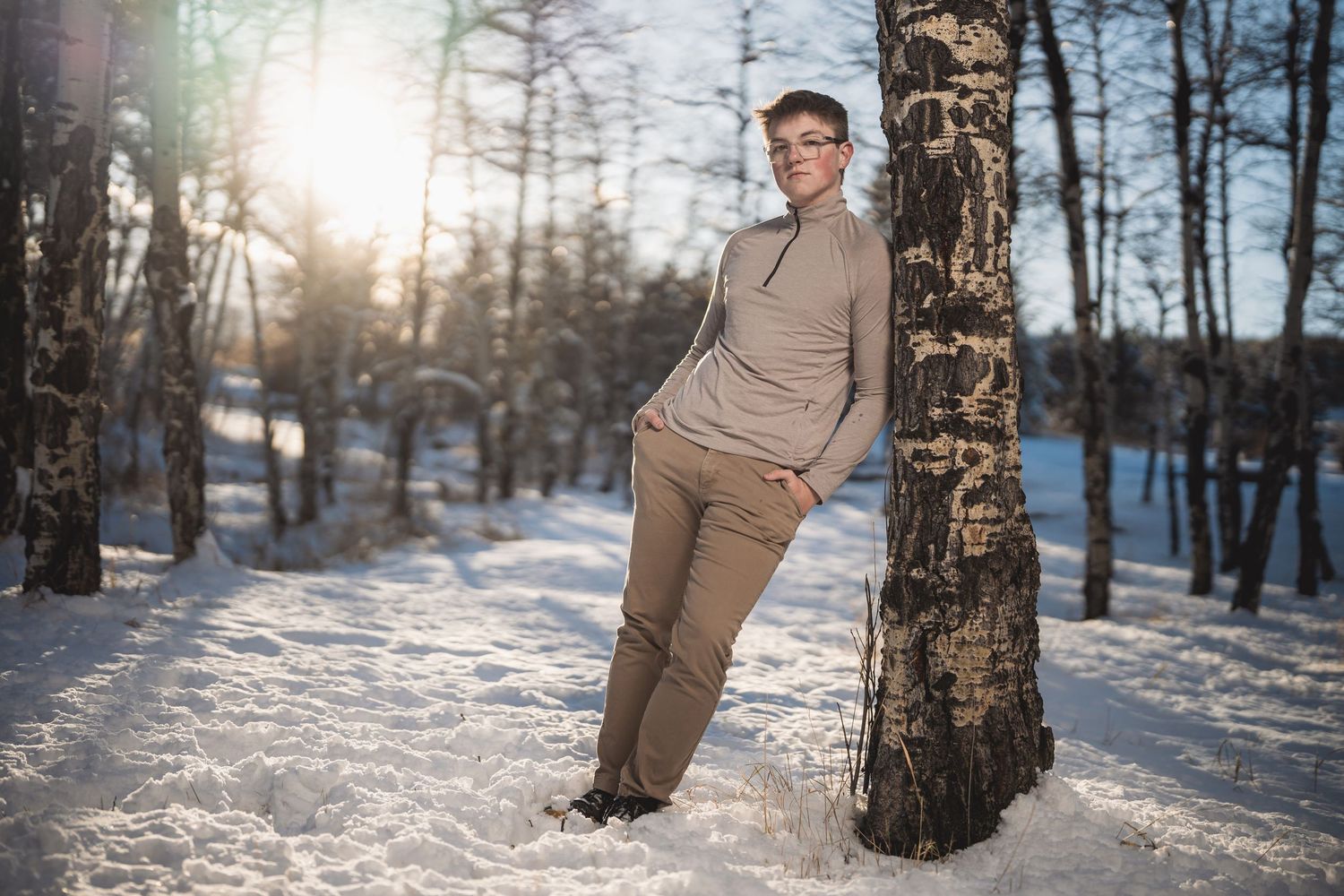 Person leaning against a birch tree in a snowy winter forest, wearing beige outfit, sunlight filtering through trees.