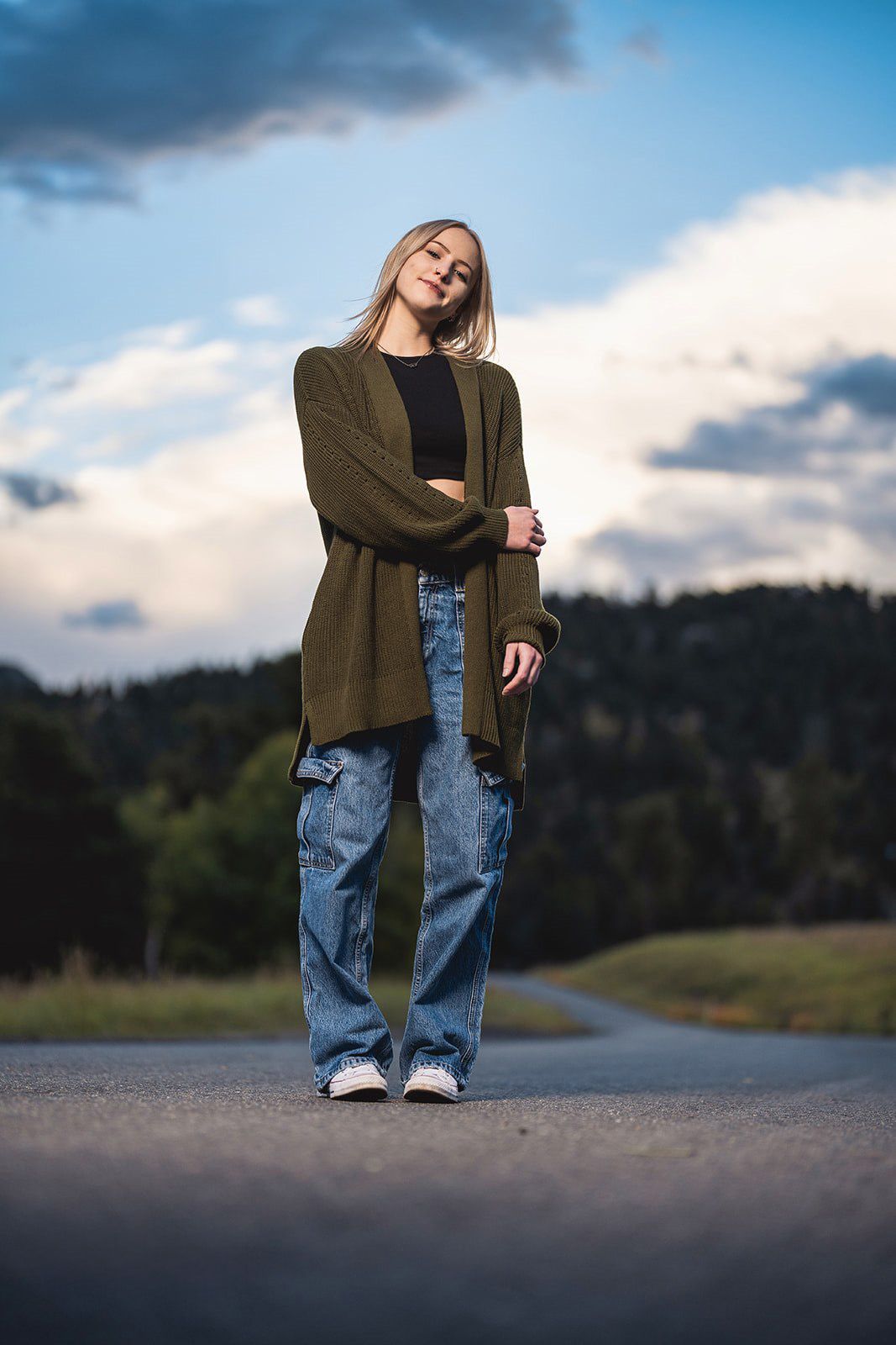 Blonde woman in olive cardigan and baggy jeans posing on a road with dramatic cloudy mountain backdrop.