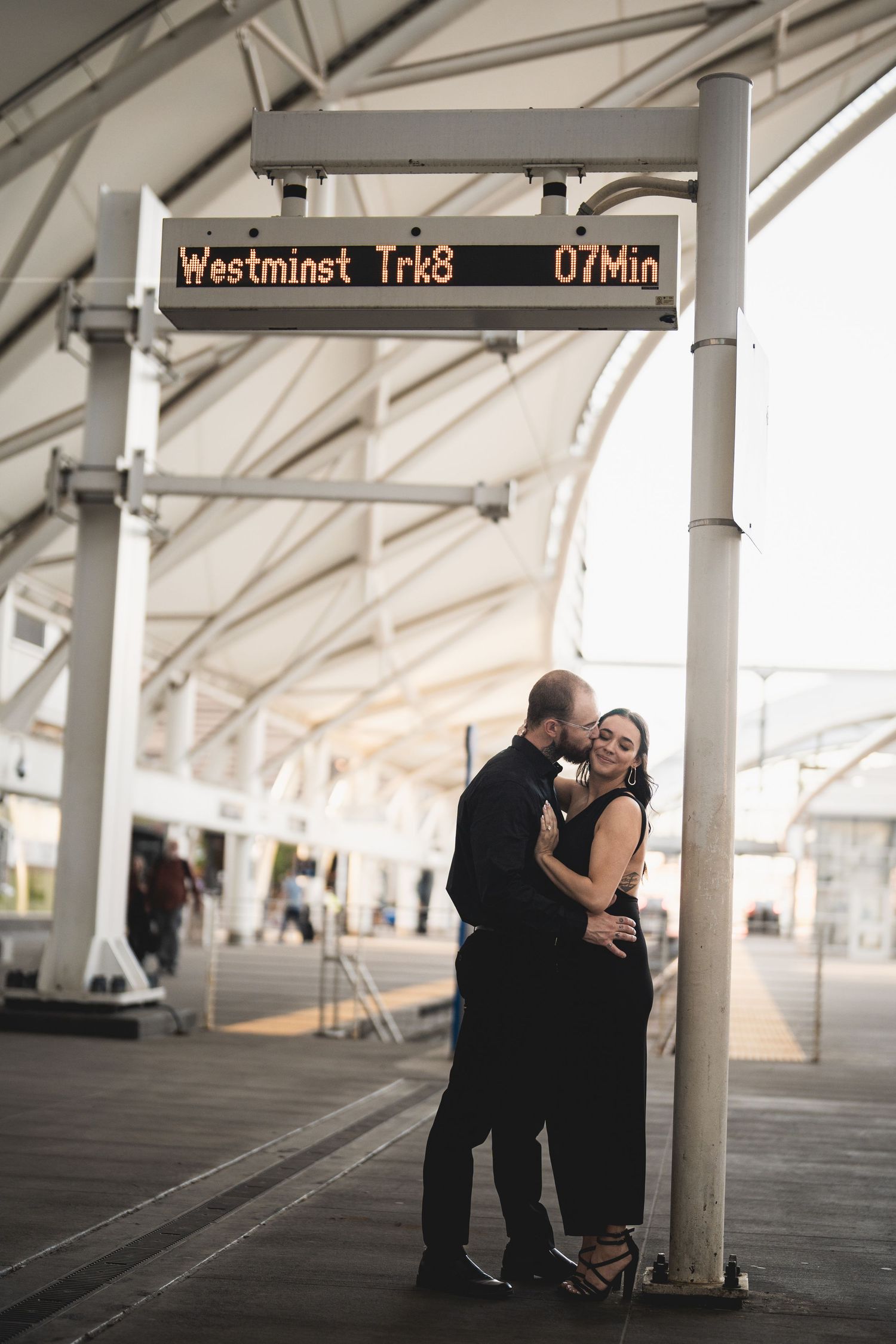 Couple in black attire embracing romantically under a train departure sign at Westminster station platform.