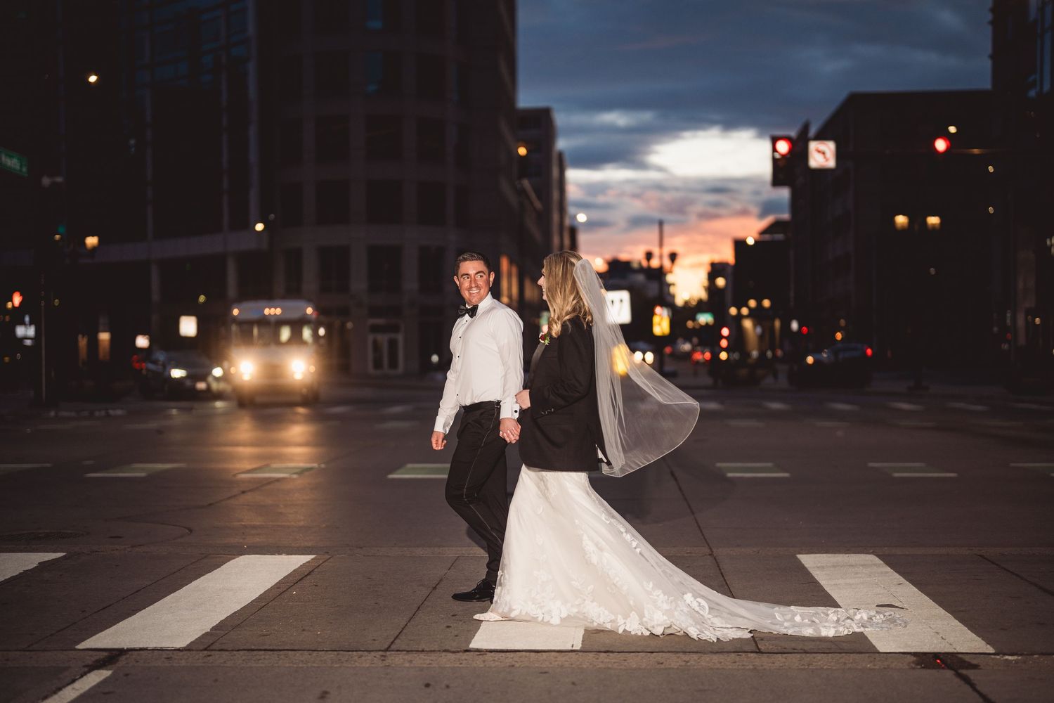 Bride and groom holding hands crossing a city street at dusk in wedding attire, with a stunning sunset skyline backdrop.
