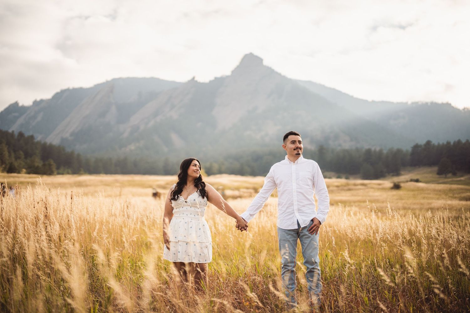 Couple holding hands in golden meadow with Rocky Mountain Flatirons backdrop during sunset engagement photo session.