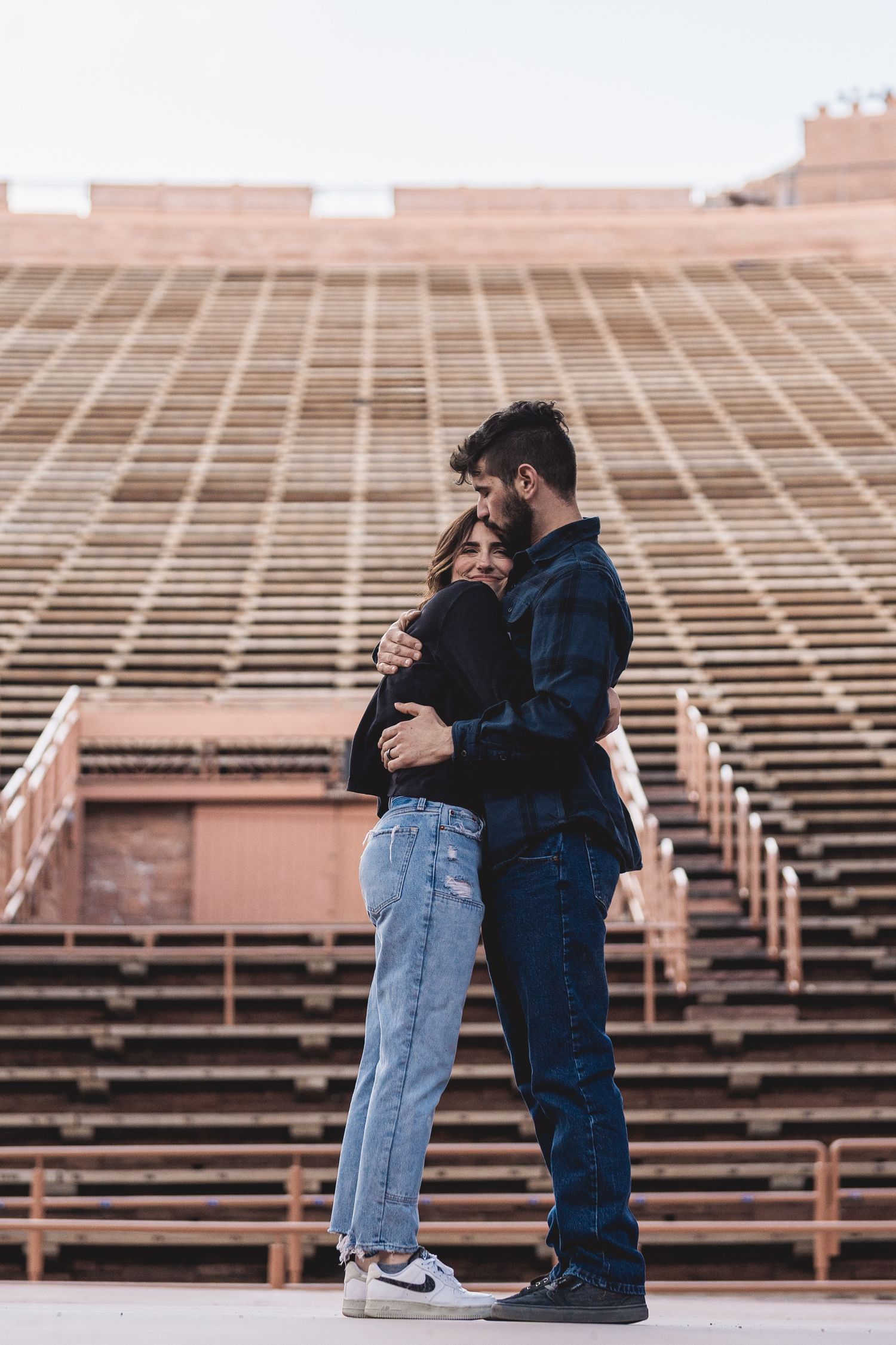 Couple sharing a tender embrace in front of empty stadium bleachers in natural light.