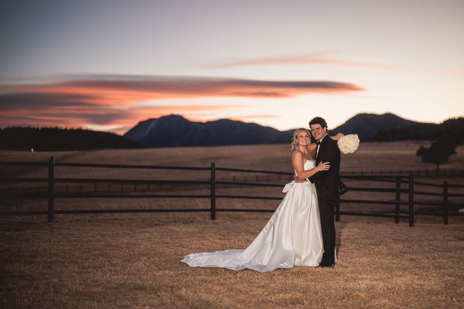 Bride and groom embrace at sunset with mountain backdrop and wooden fence at outdoor ranch wedding venue.