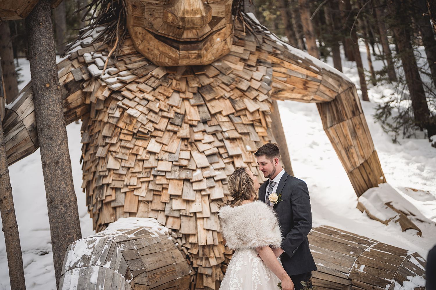 Bride and groom share a romantic moment in front of a large wooden troll sculpture in a snowy winter forest.