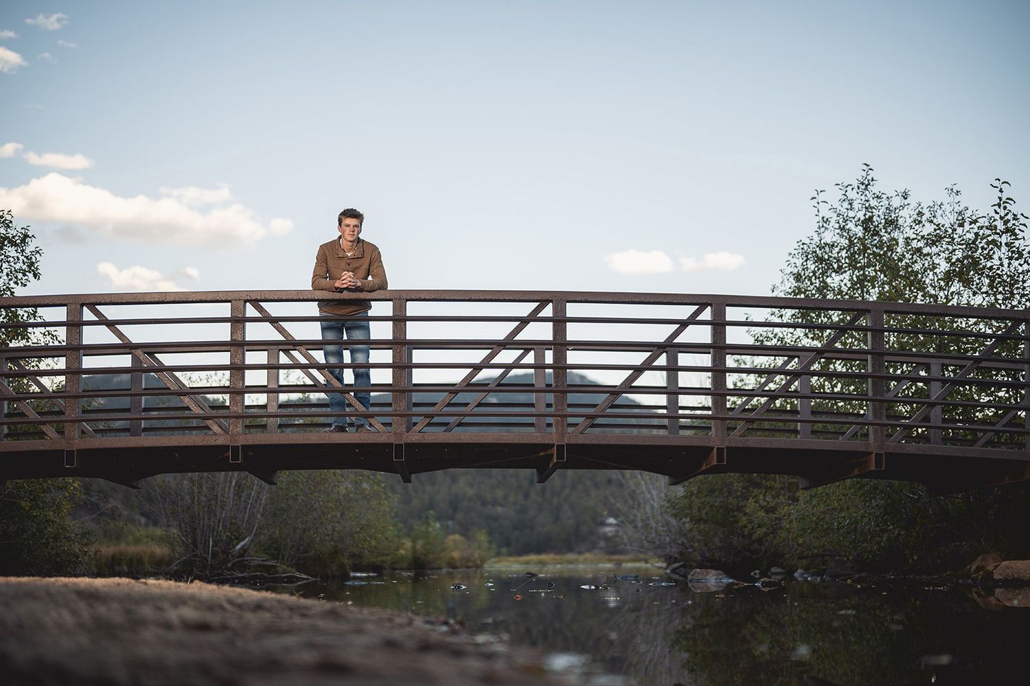 Young man standing on wooden bridge over river in scenic outdoor setting.