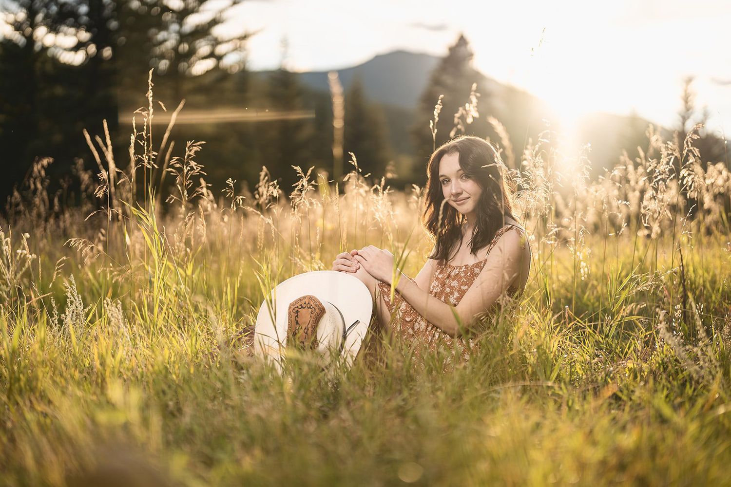 Young woman in floral dress sitting in sunlit meadow holding a white cowboy hat.