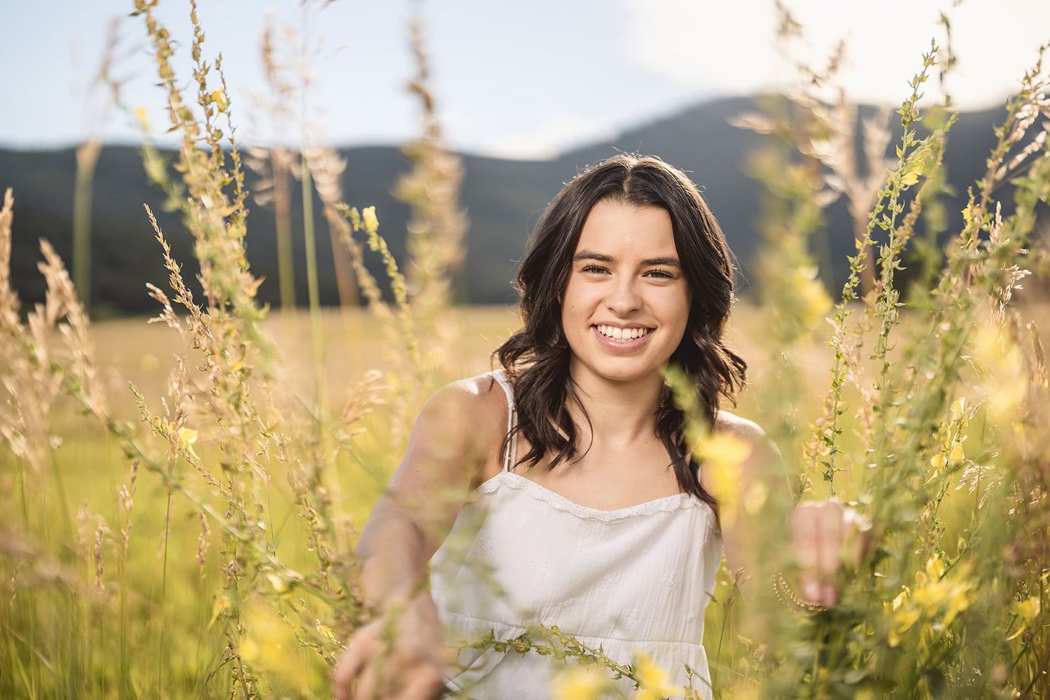 Smiling young woman in white dress standing among tall yellow wildflowers in a sunny mountain meadow.
