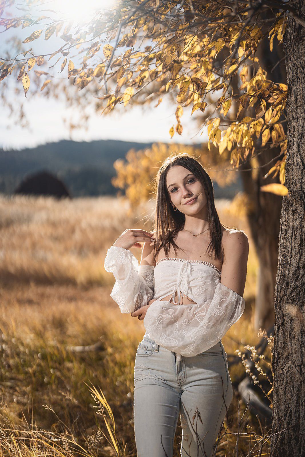 Young brunette woman in white off-shoulder top and jeans leaning against autumn tree.