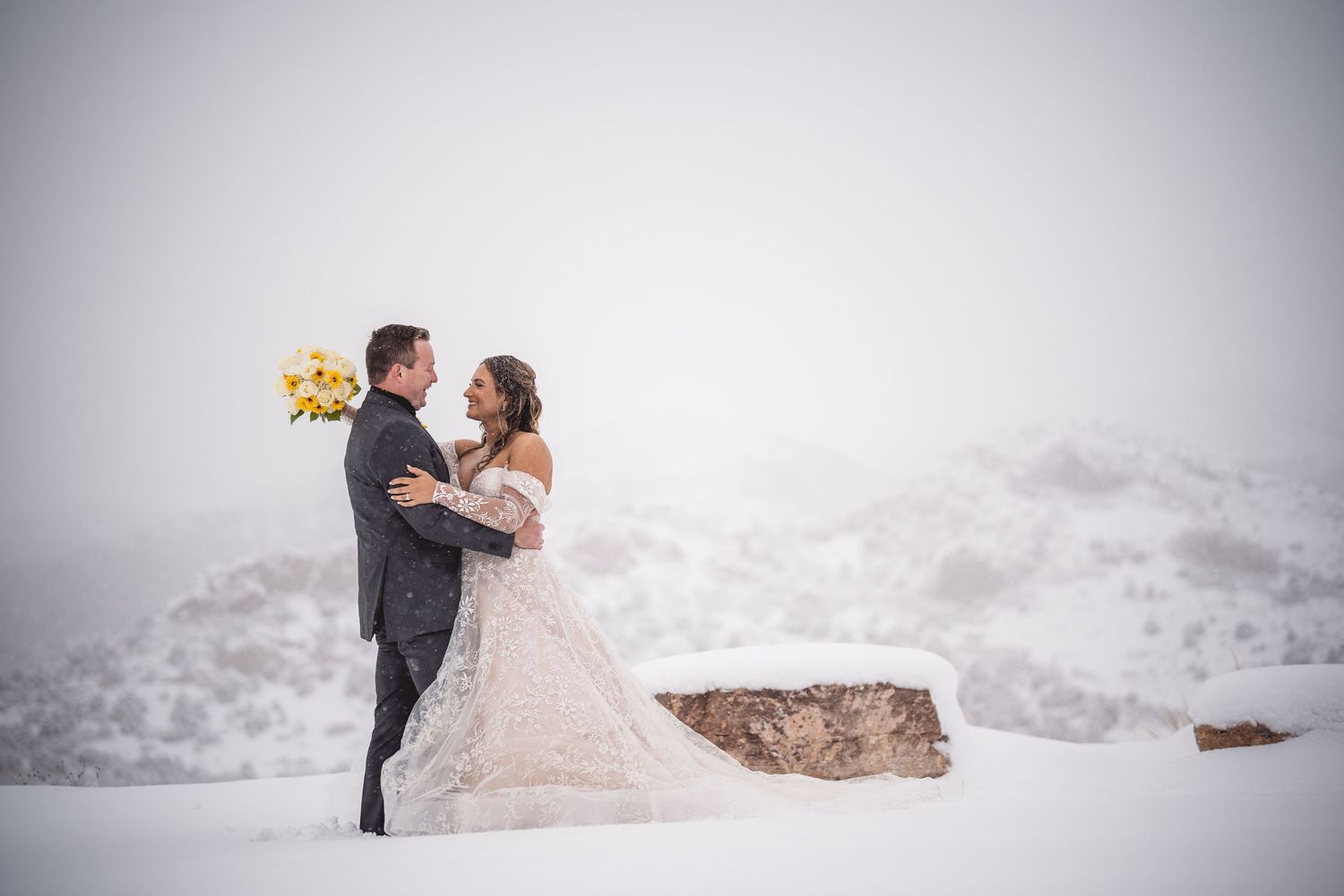 A couple embraces in a snowy winter landscape, the bride holding a yellow bouquet in a lace gown.