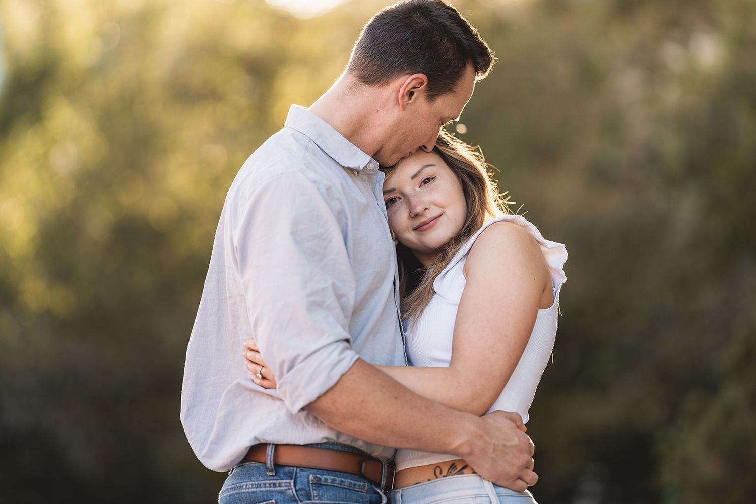 Couple embracing outdoors at golden hour, woman resting head on man's chest in a romantic engagement photo.