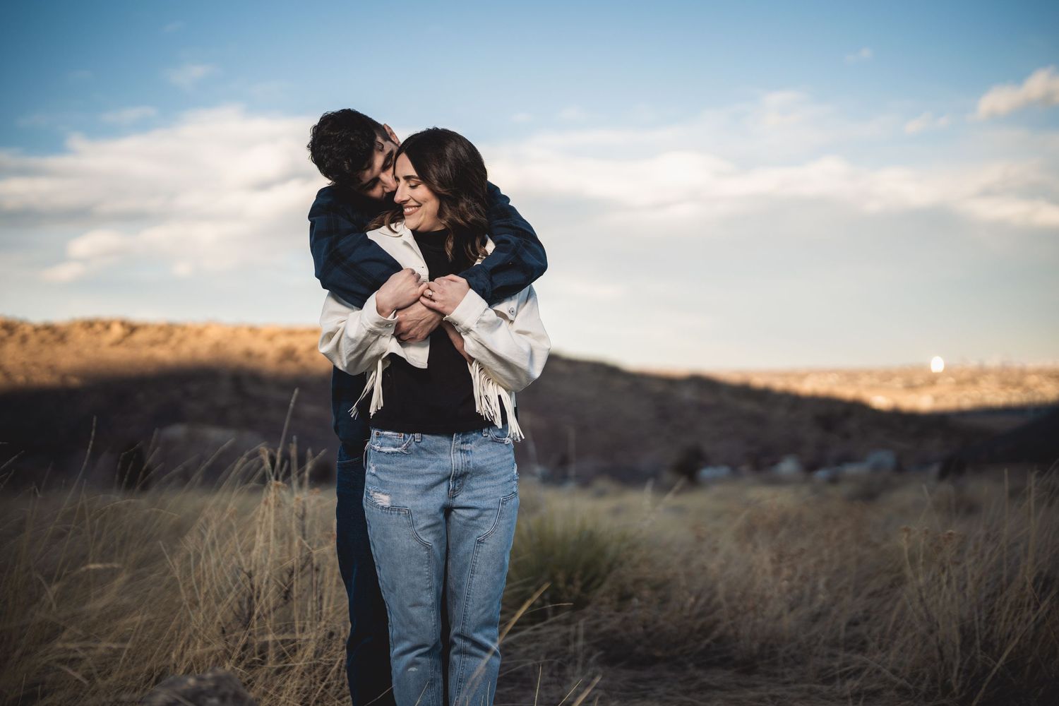 A couple embraces warmly in a golden field under a cloudy sky, sharing a tender romantic moment outdoors.