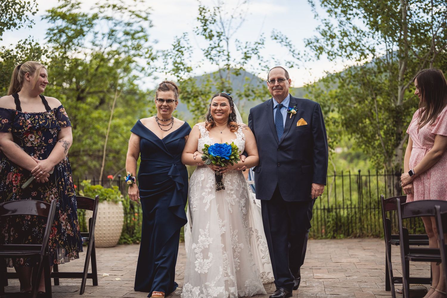 A wedding ceremony in progress at an outdoor mountain venue with guests seated on wooden chairs.