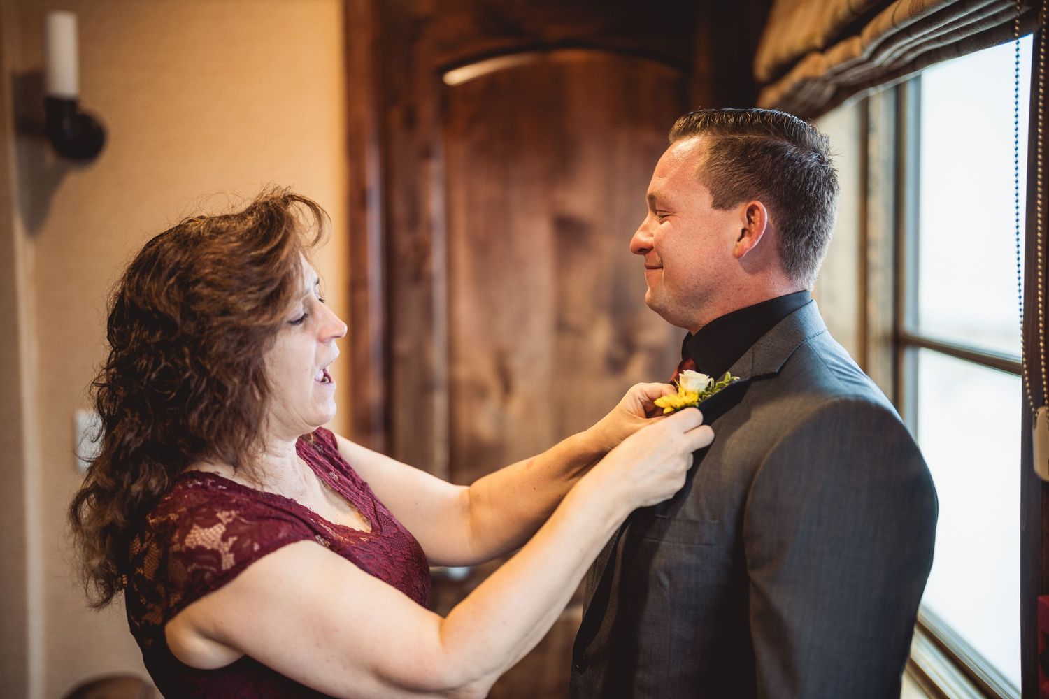 Someone helps adjust a boutonniere on formal wedding attire near a window.