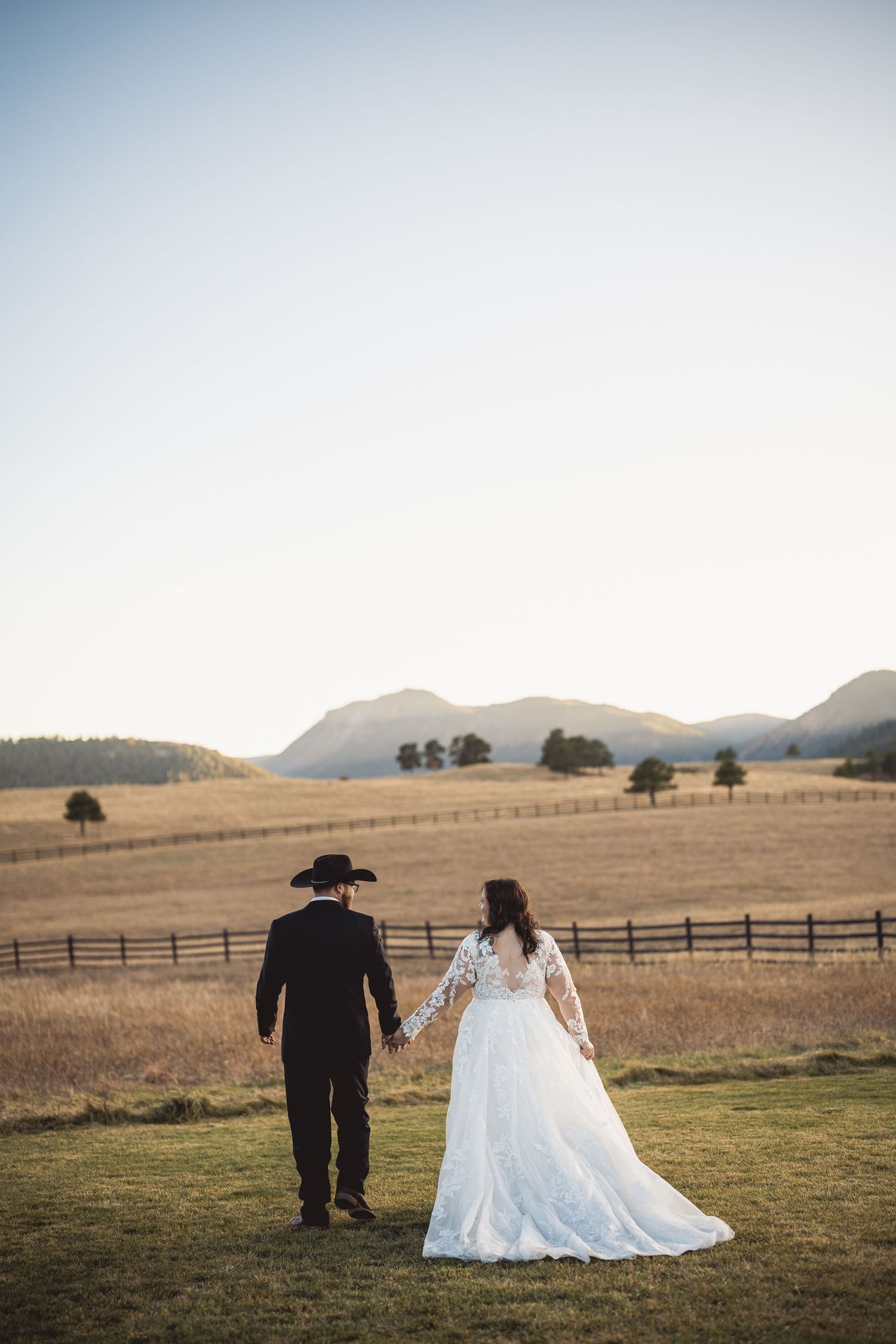 Wedding couple standing in rustic field with mountains and wooden fence in background at sunset.