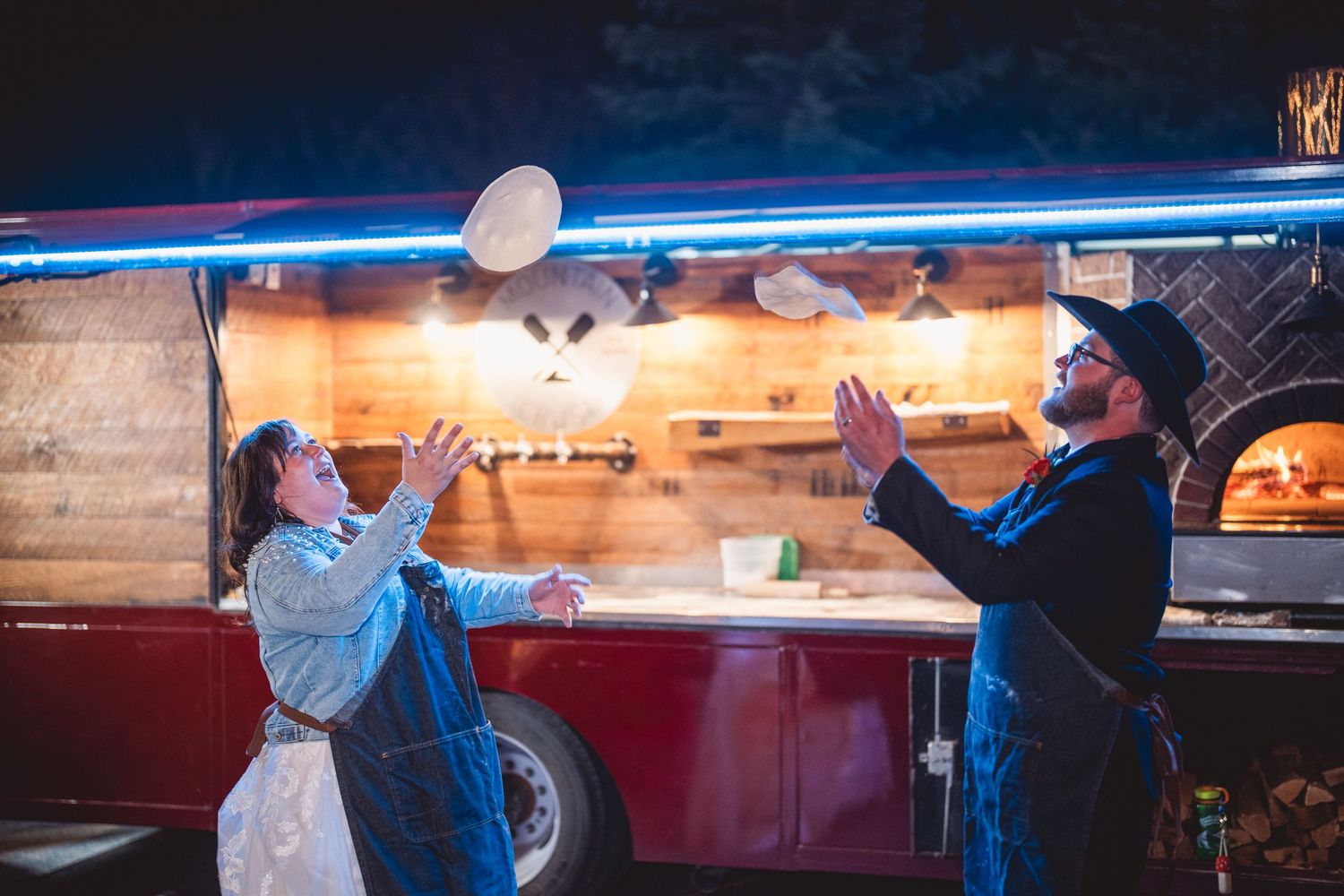 Wedding couple playfully tossing pizza dough at food truck at night.