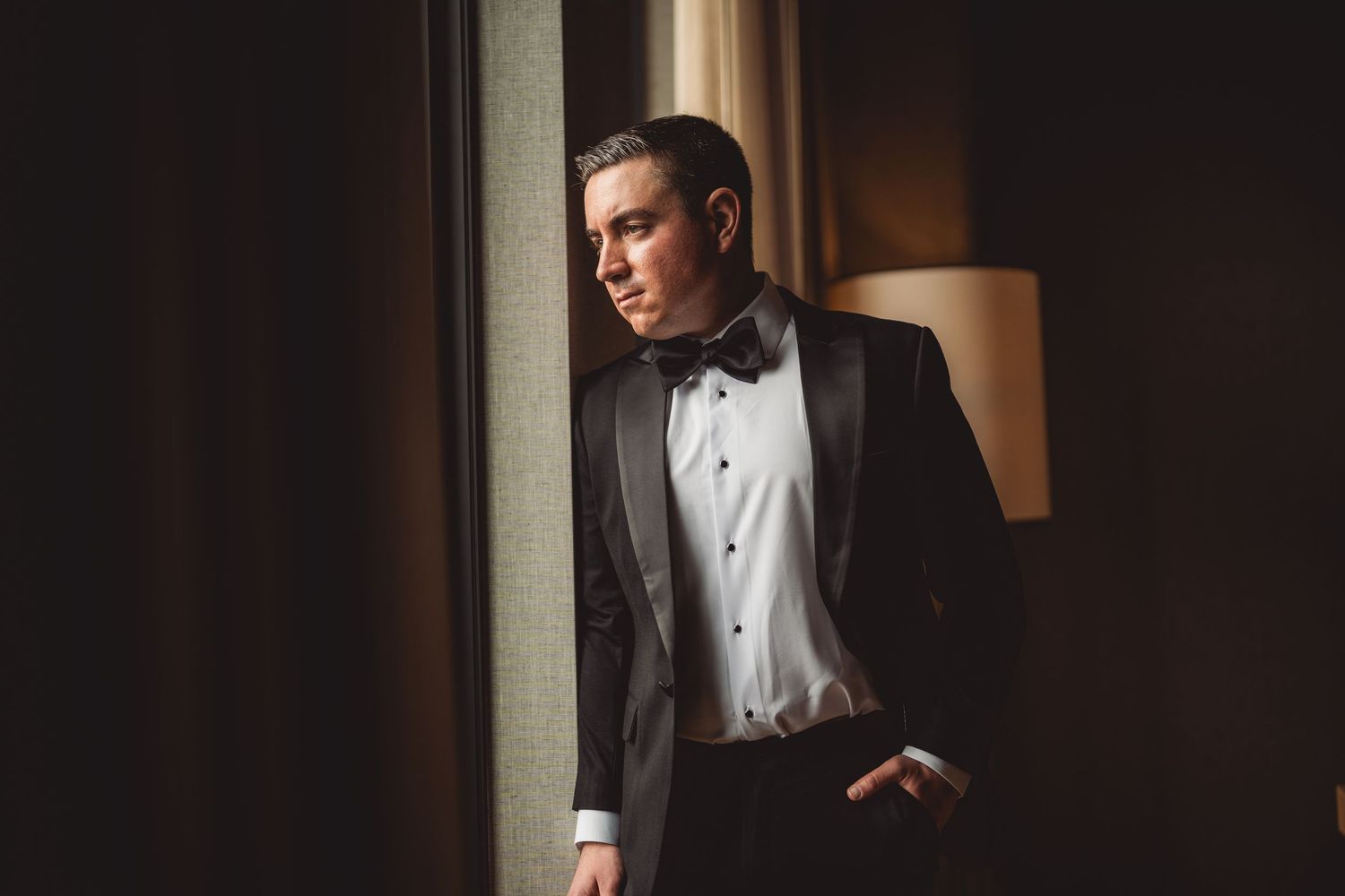 Person in classic tuxedo and bow tie standing near window in moody lighting.