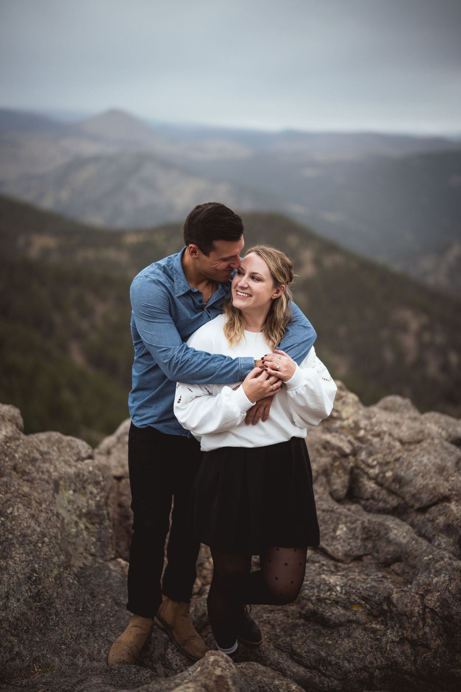 A romantic couple shares an intimate moment while sitting on rocky mountain overlook during sunset.