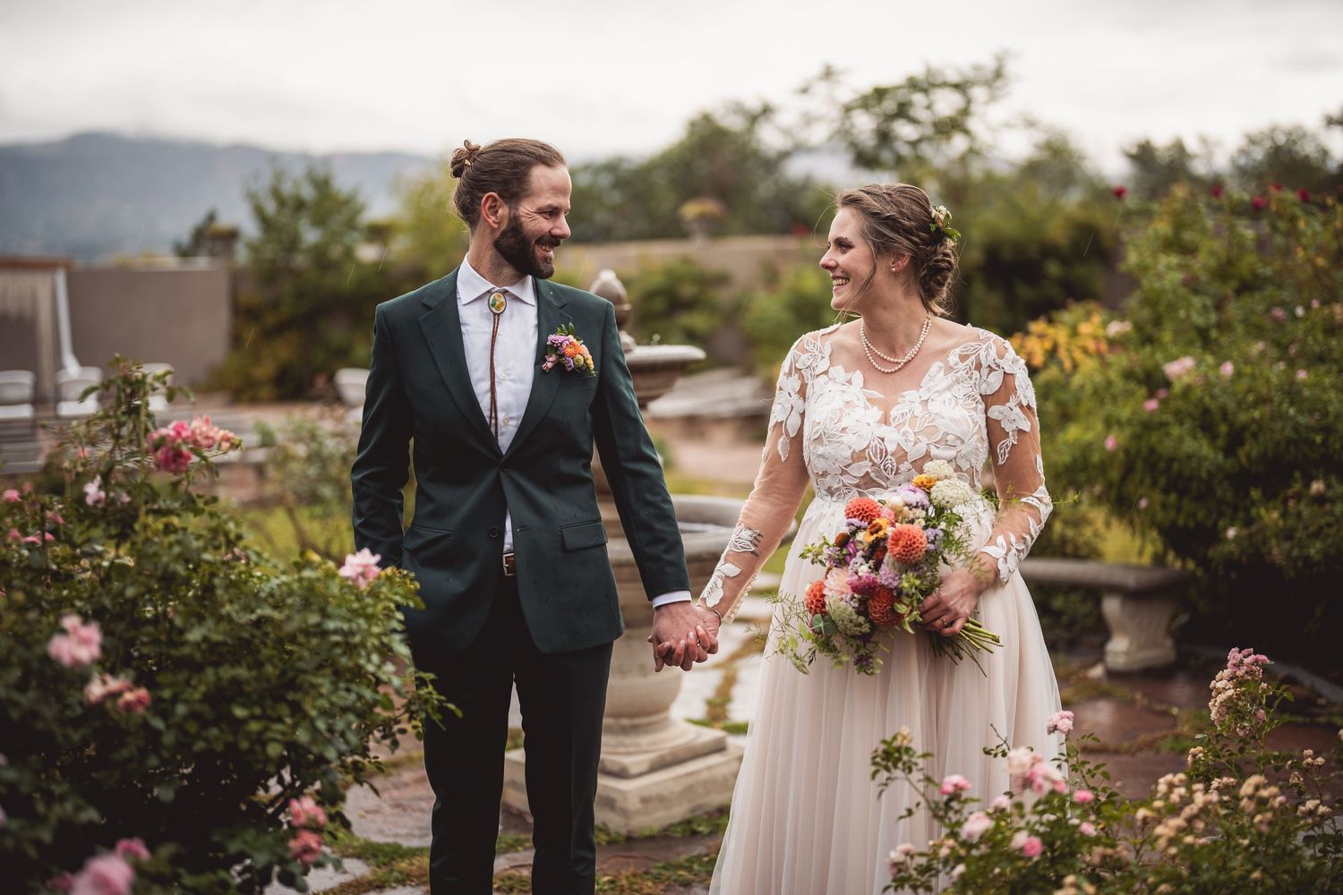 Wedding couple walking through garden path surrounded by pink roses and mountain views.