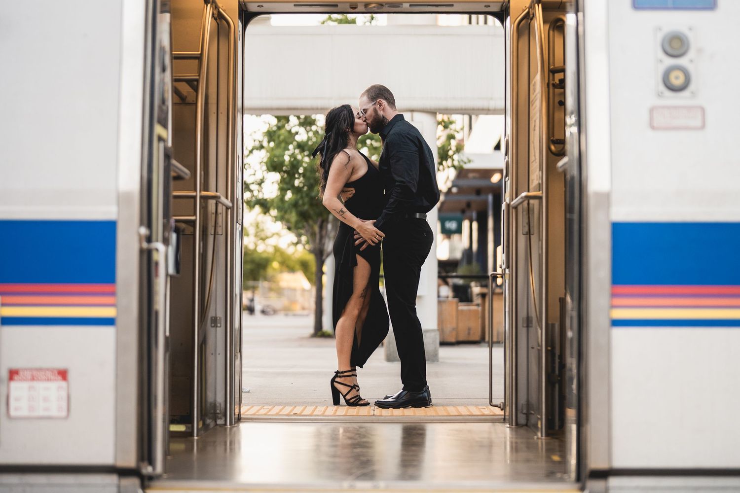 Couple sharing a romantic kiss in the doorway of a train, woman in black dress and heels, man in suit.
