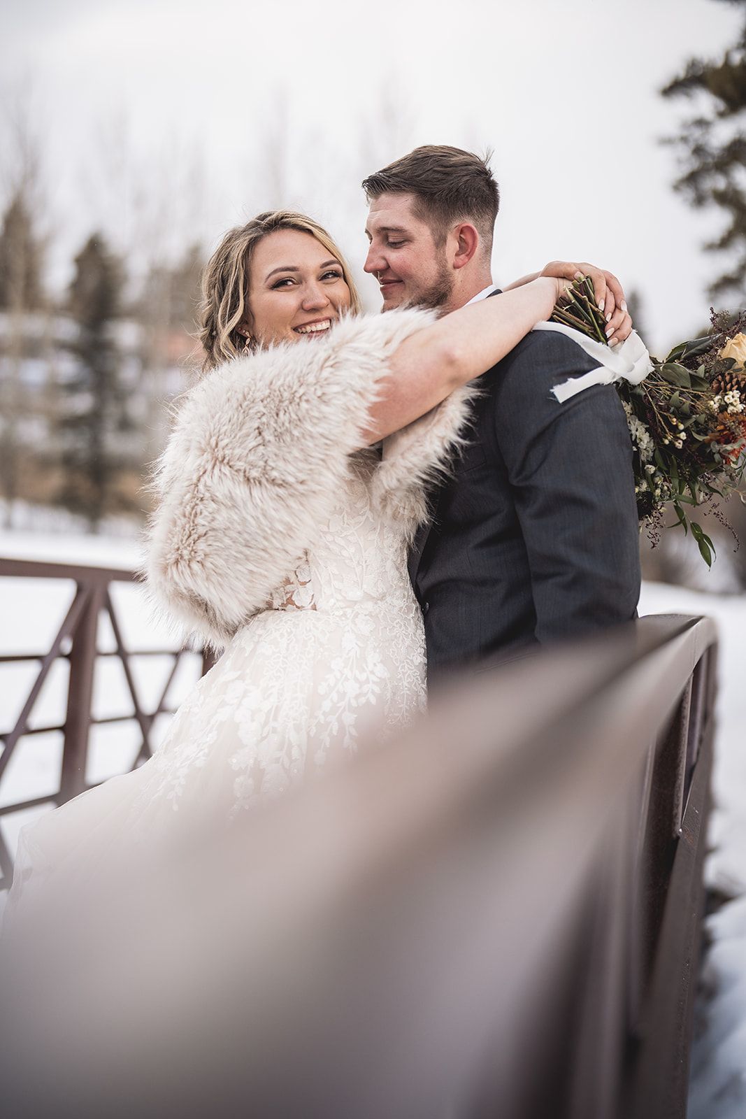 Bride in white fur wrap and groom in dark suit embrace on snowy winter bridge holding a bouquet.