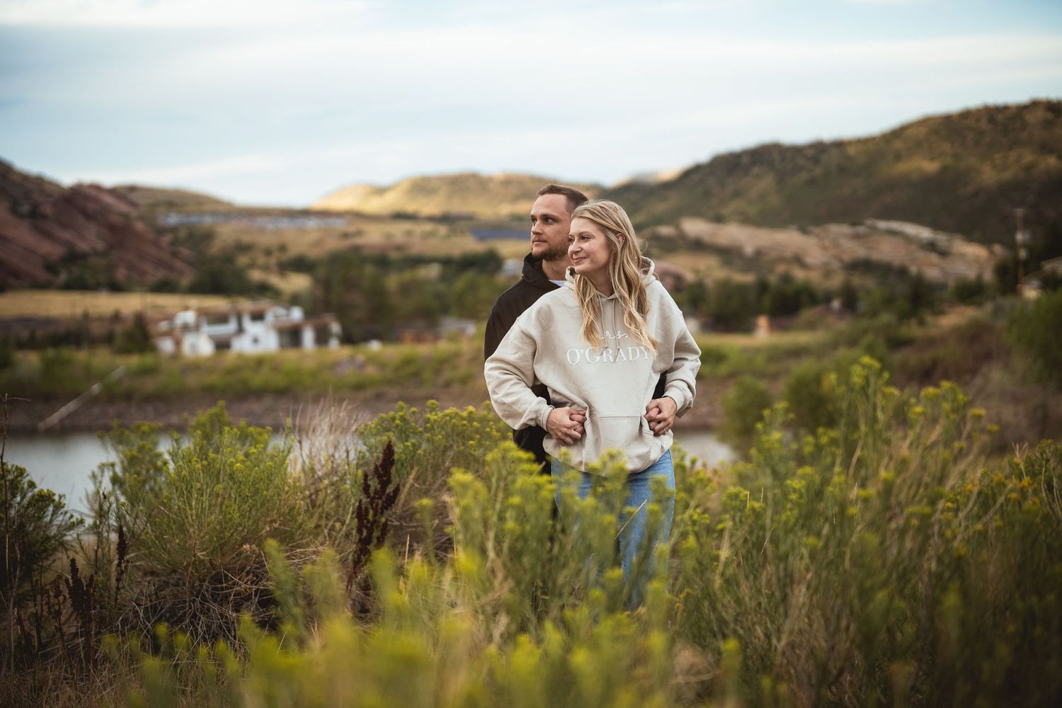 A couple smiles together among green shrubs with rolling hills and a lake in the background outdoors.