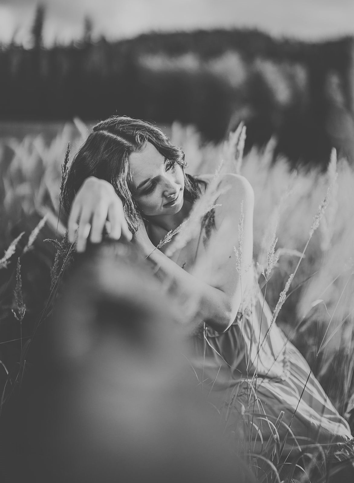 Black and white photo of a young woman sitting peacefully among tall wild grass in a serene outdoor field.