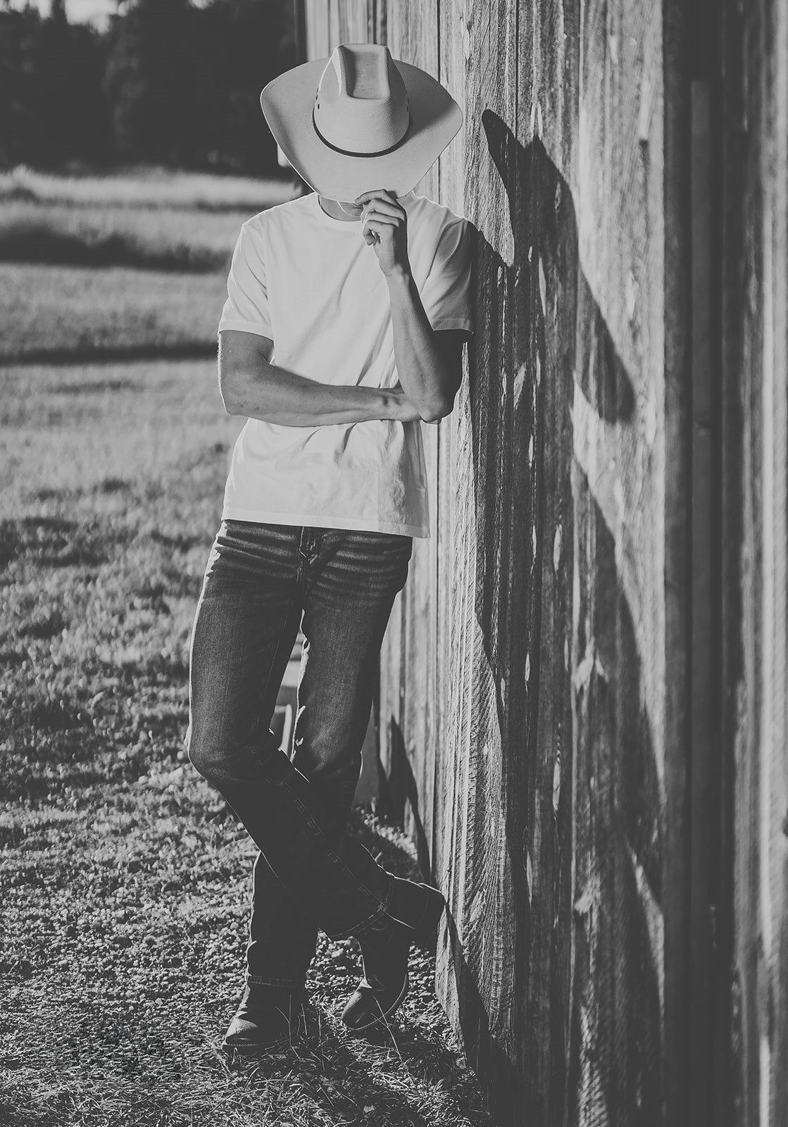 Young man in cowboy hat, white tee, jeans, and boots leaning against rustic barn wall in black and white photo.