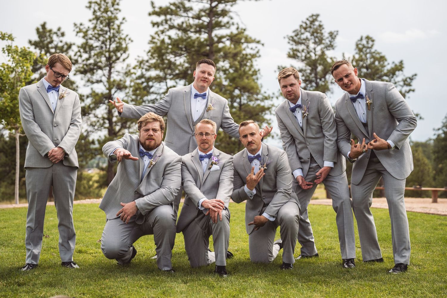 Groomsmen in gray suits and blue bow ties pose playfully outdoors on grass with trees in the background.