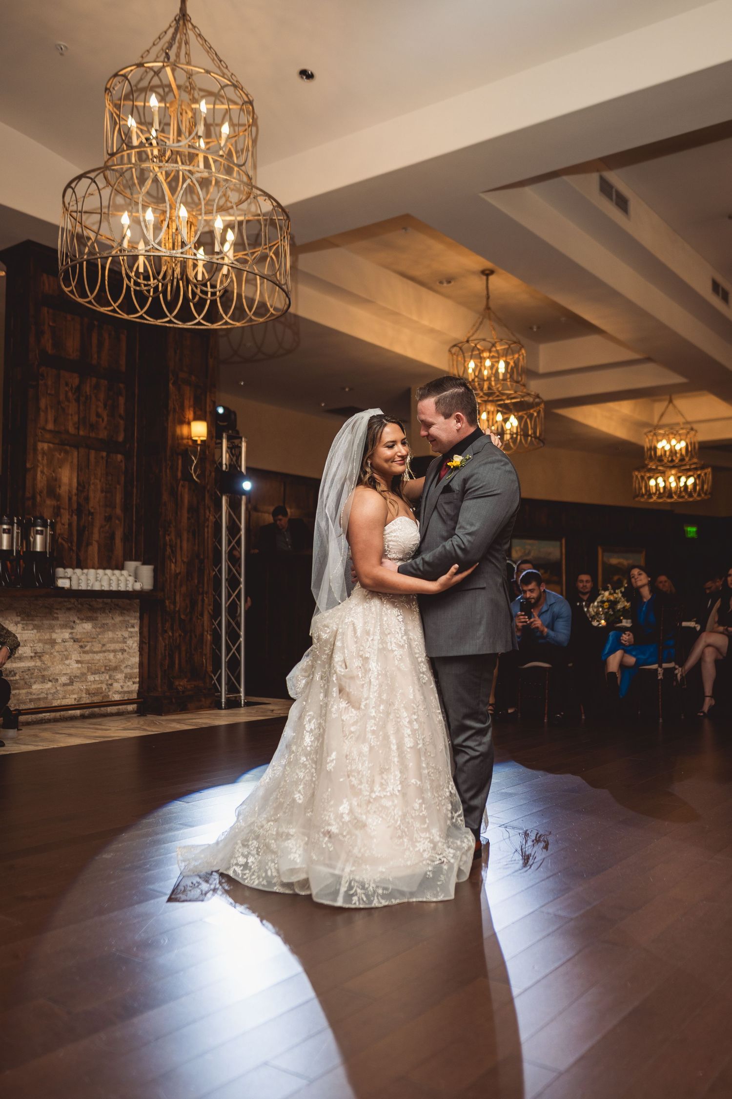 Bride and groom share their first dance under elegant gold chandeliers in a warmly lit wedding reception venue.