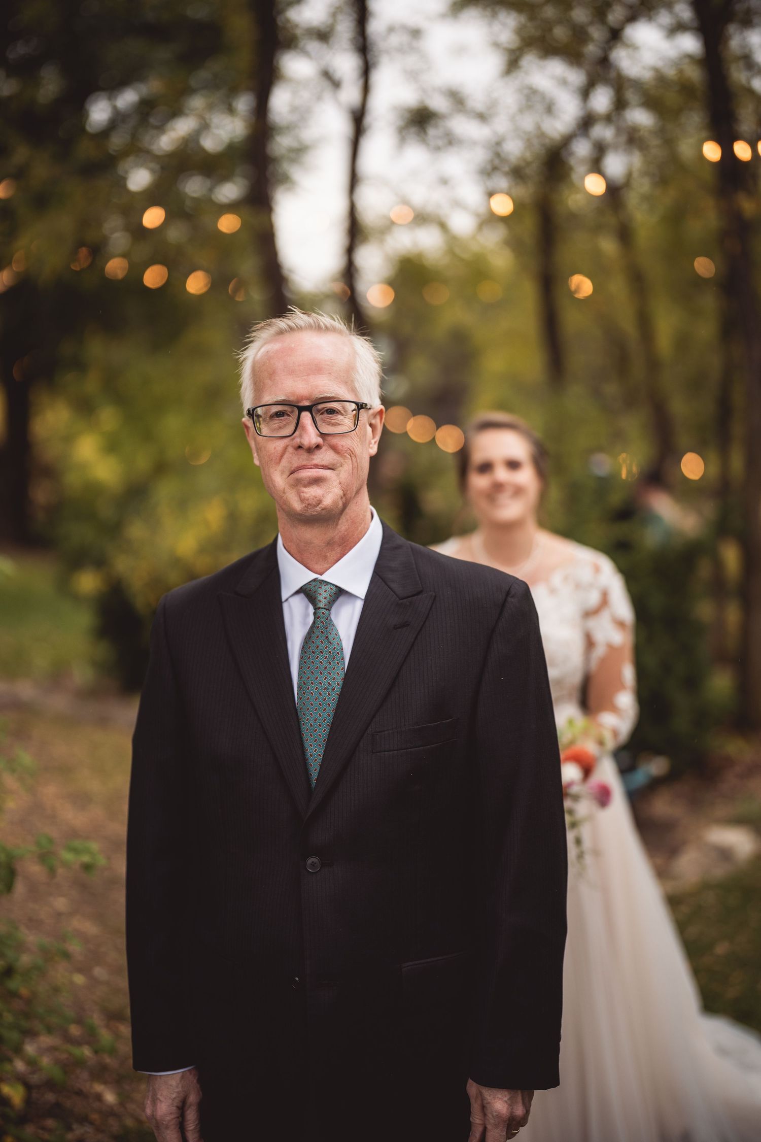 A formal wedding portrait taken outdoors with string lights and fall trees in the background.
