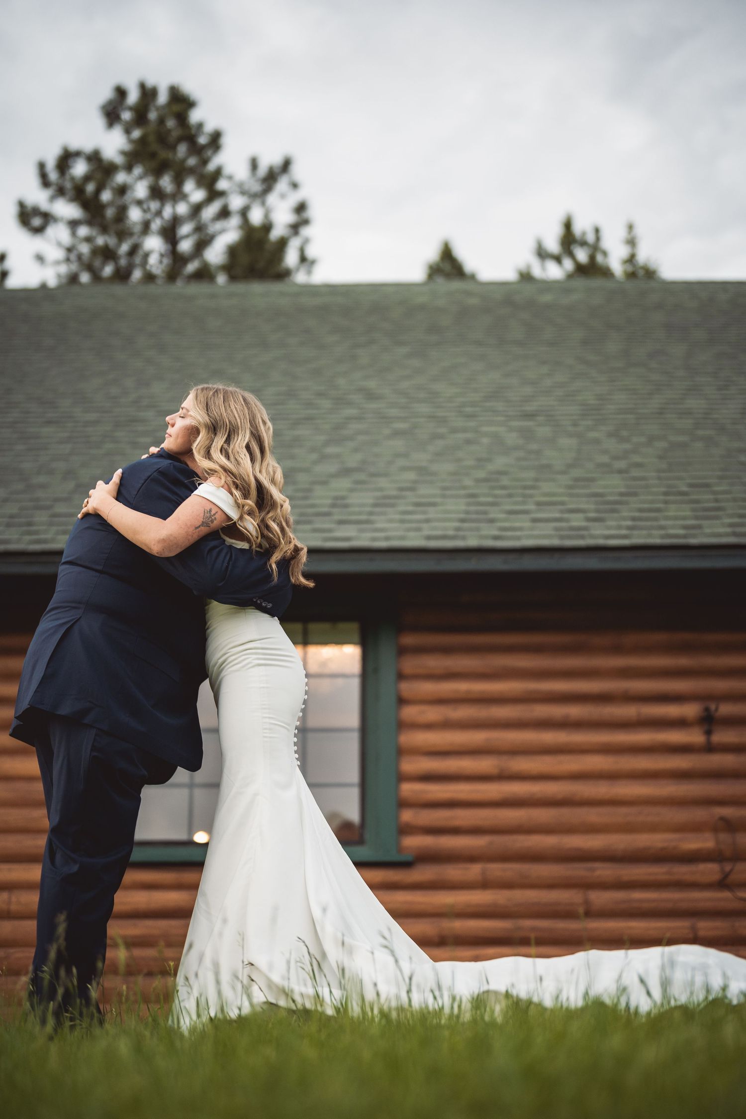 Wedding couple embraces outside rustic log cabin with green grass and grey roof.