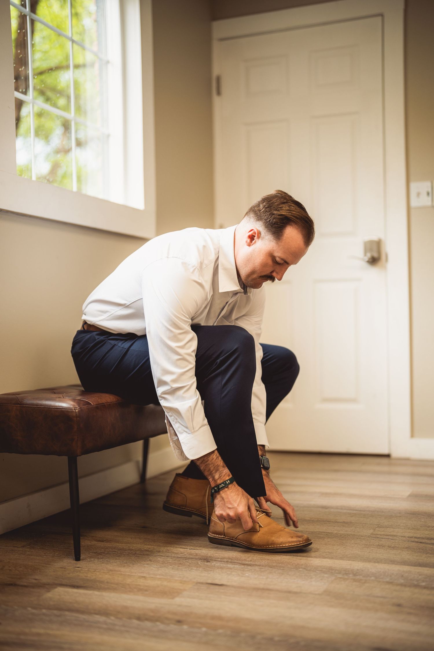 Person in formal attire adjusting brown leather dress shoes on wooden floor.