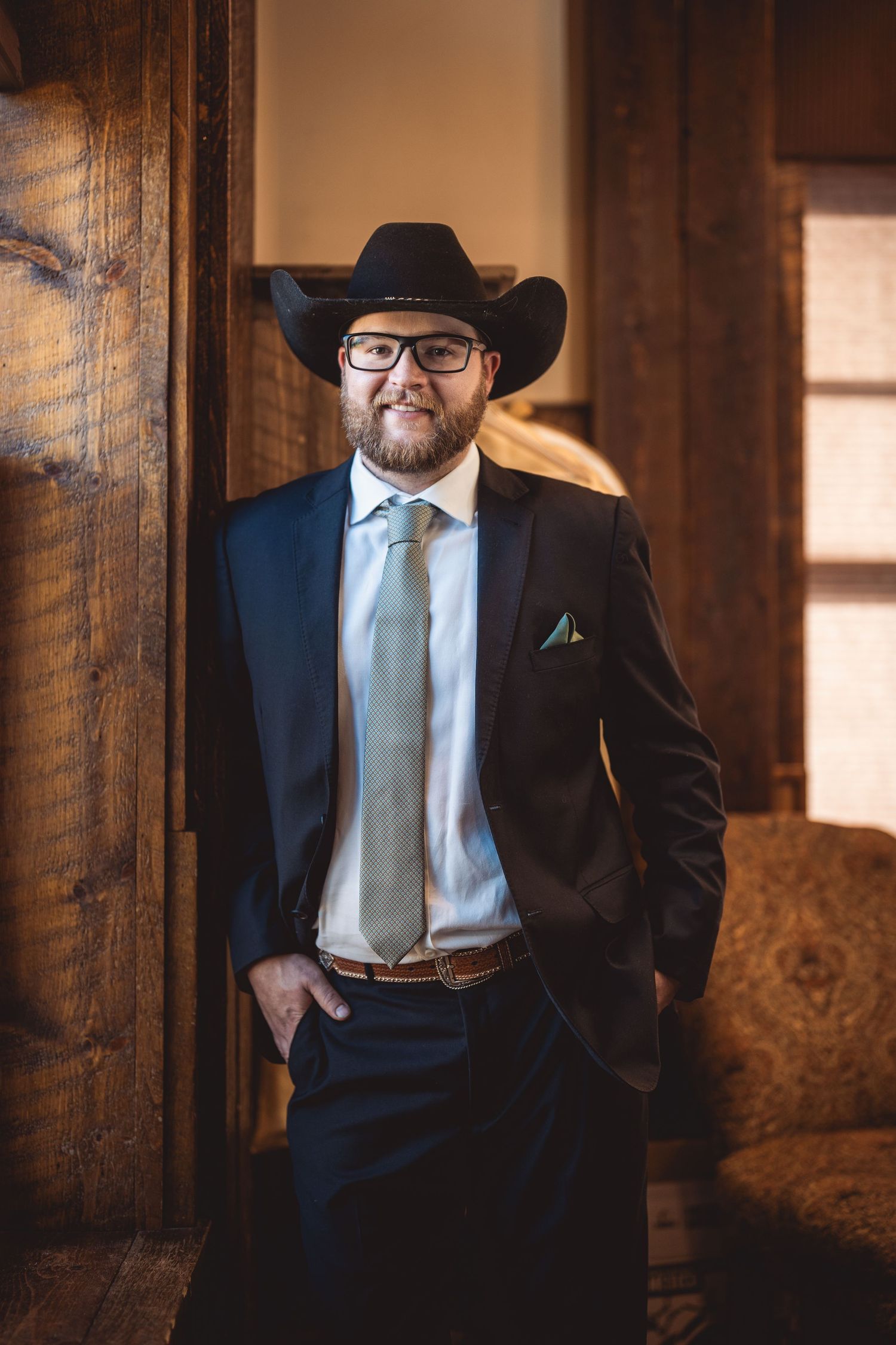 Western-style groom in black cowboy hat and suit poses against rustic wooden interior walls.