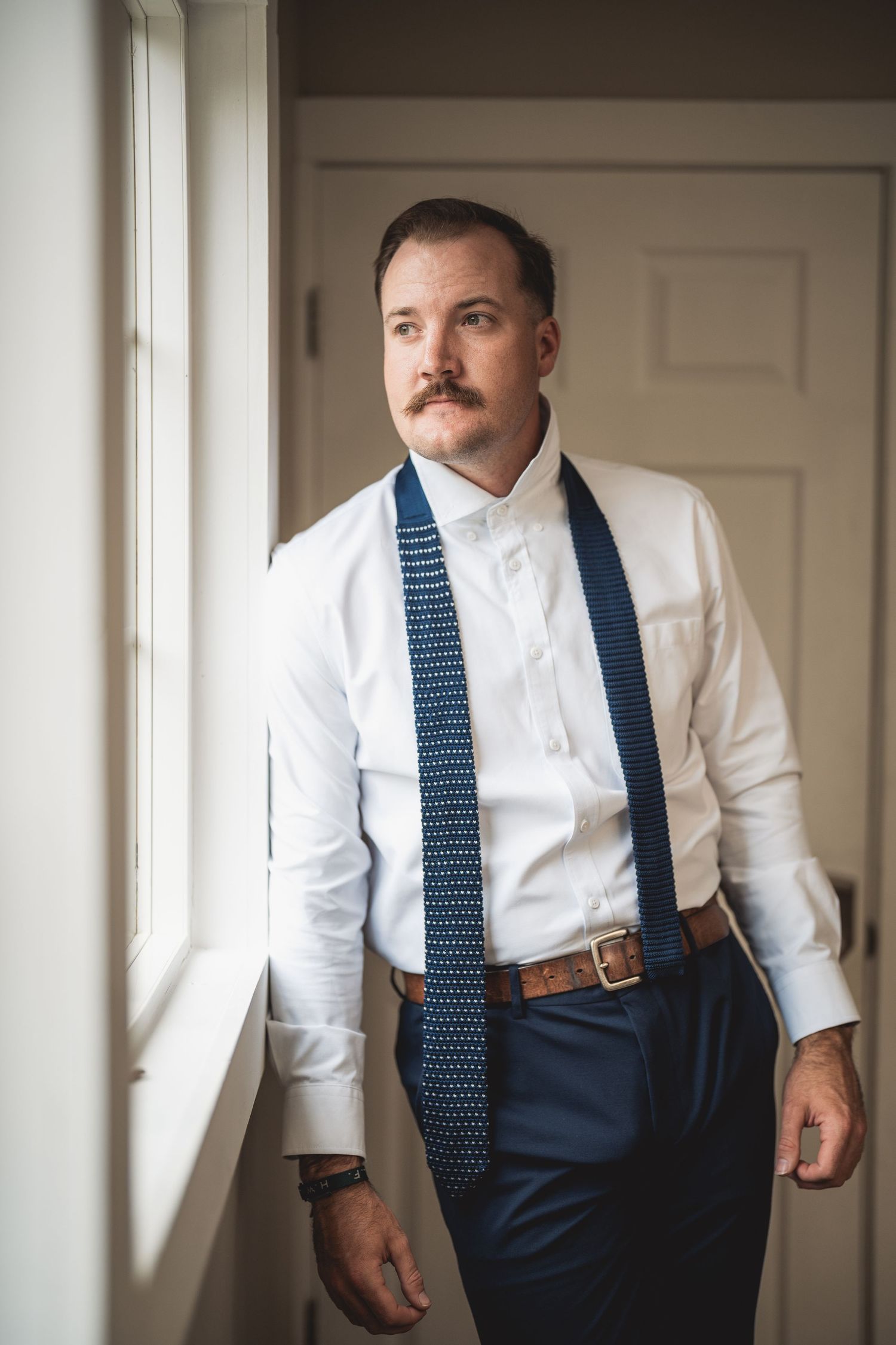 A man in formal attire adjusts his navy blue polka dot tie while getting dressed.