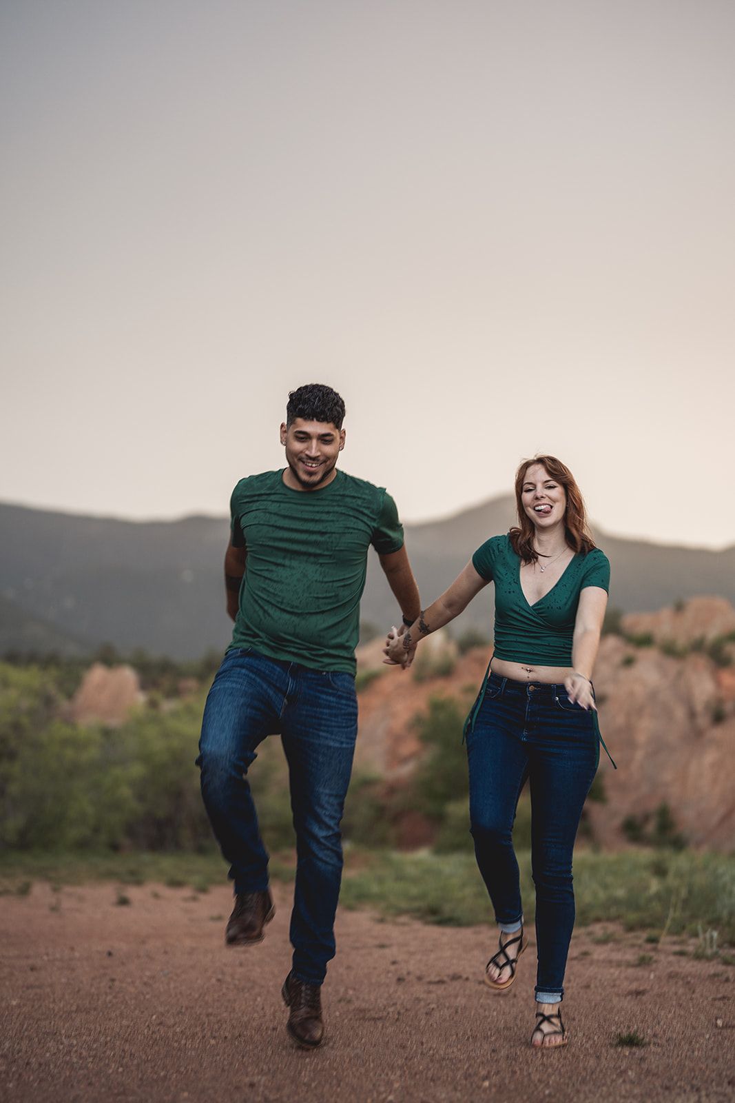 Joyful couple running and laughing together on a dirt path with mountains in the background.