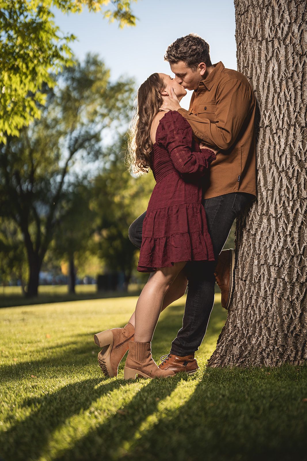 Couple in burgundy and brown outfits shares playful moments near tree trunk in sunny park.