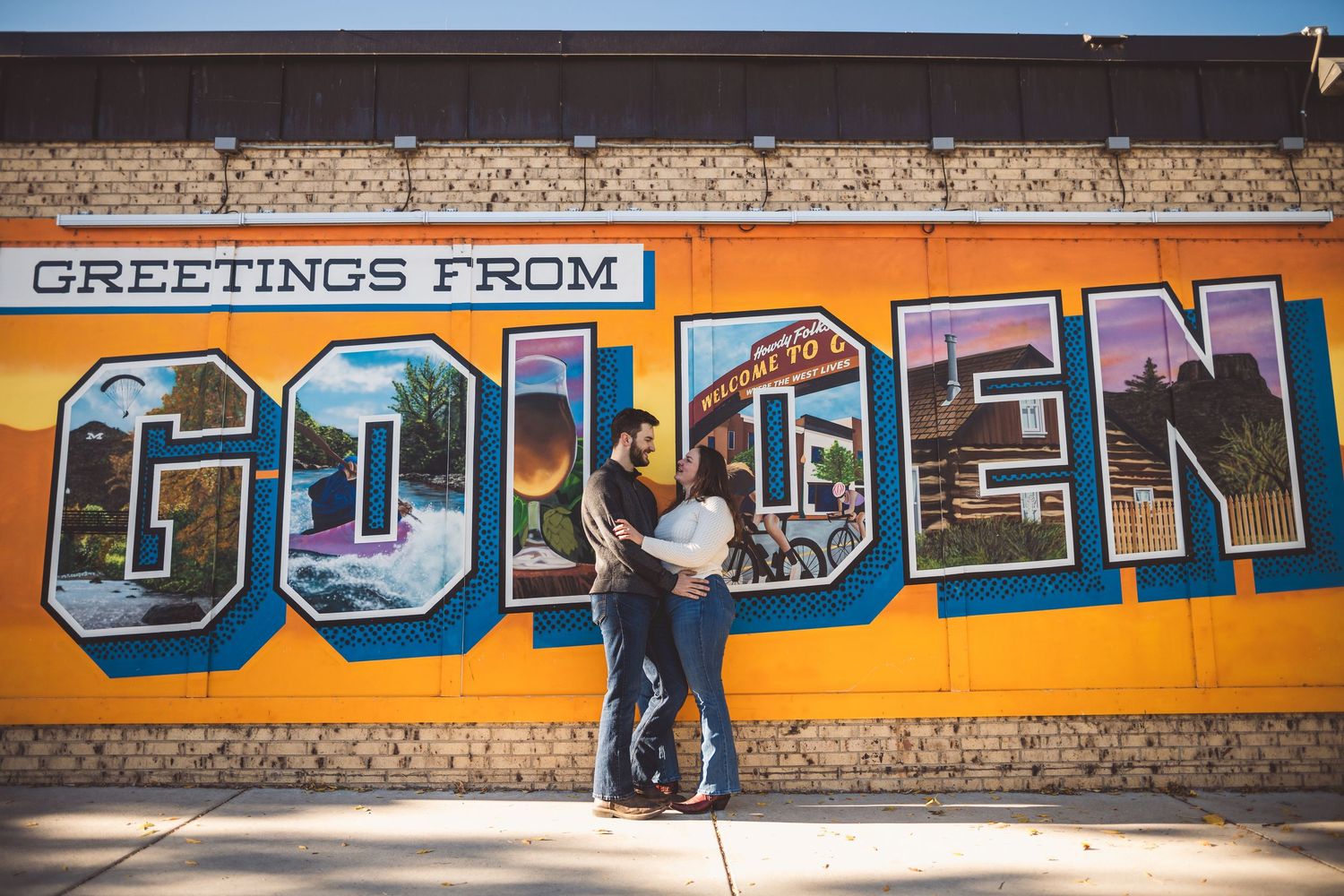 Couple embracing in front of colorful Greetings From Golden mural on brick wall.